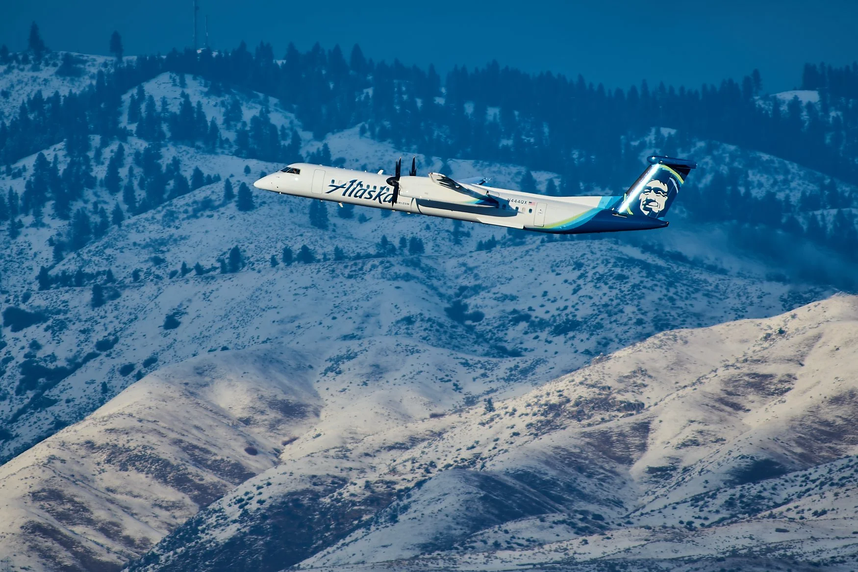 An Alaska Airlines Bombardier Dash 8 turboprop airplane flying over snow-covered mountains with a forest in the background.