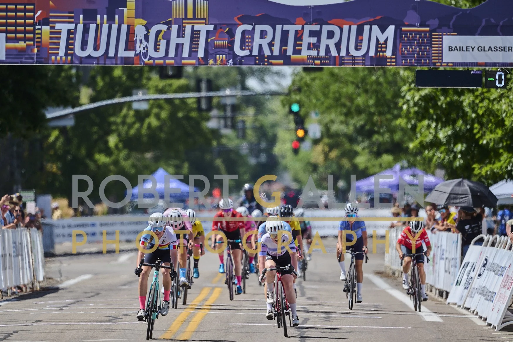 Cyclists racing in a cycling race event, with the finish line and spectators on sides, under a banner reading 'Twilight Criterium'.