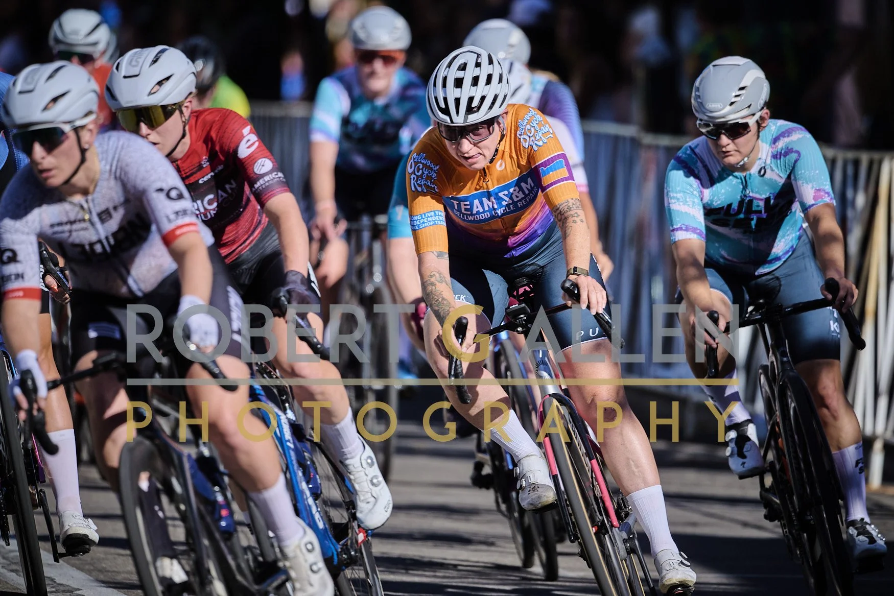 Cyclists participating in a race, wearing helmets and colorful jerseys, riding on a paved road with metal barriers on the side.
