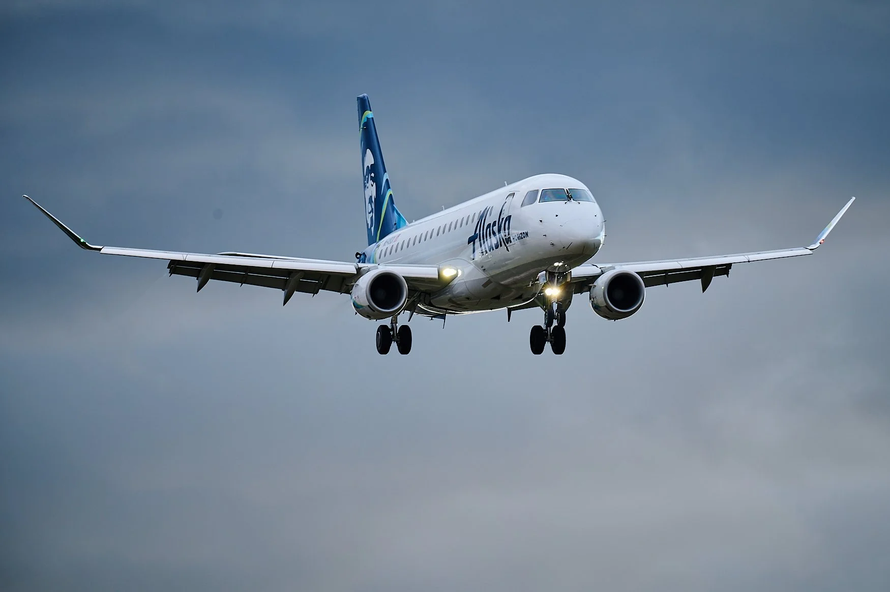 A white airplane with Alaska Airlines logo approaching for landing against a dark, cloudy sky.