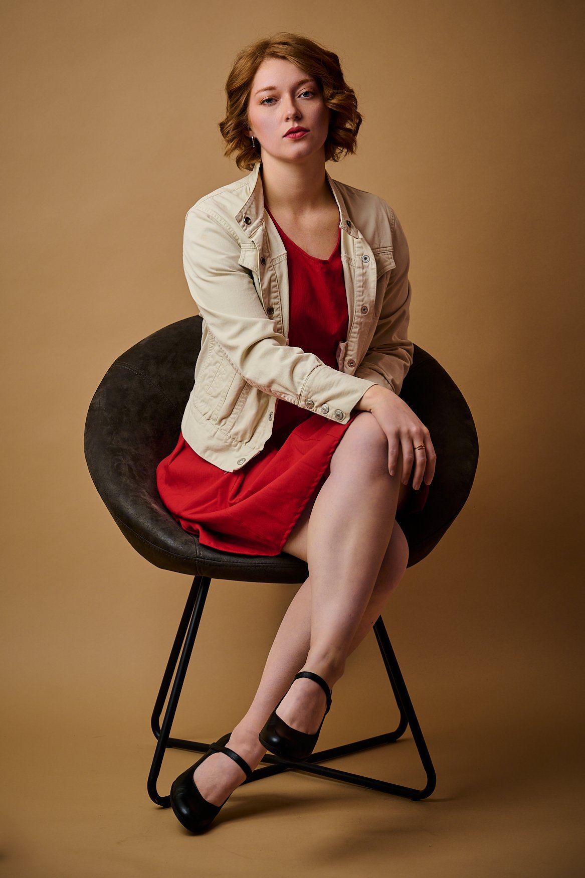 Young woman with short curly red hair sitting on a modern black chair against a beige background, wearing a red dress, beige jacket, and black shoes.