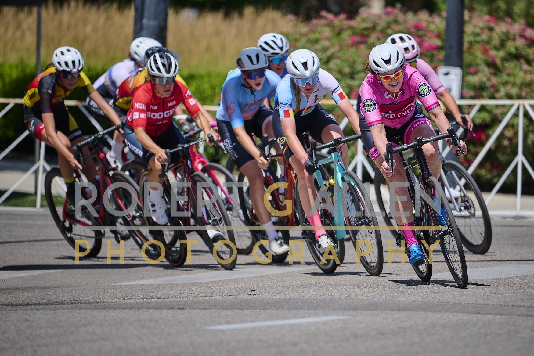 Women participating in a cycling race, wearing colorful jerseys and helmets, riding racing bicycles on a paved road with a grassy and bushy background.