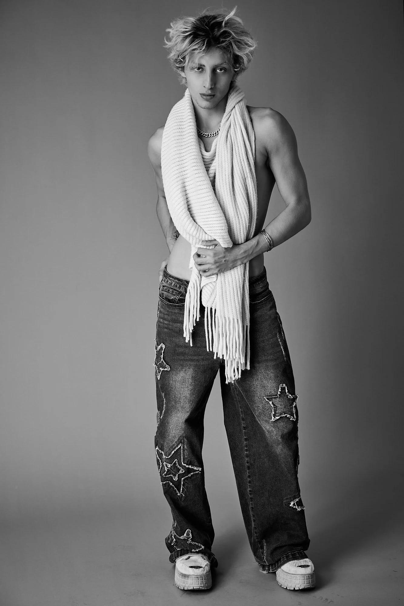 A young man with long, curly hair styled upward, wearing distressed jeans with star patches, white platform sneakers, a chunky chain necklace, and a sweater draped over his shoulders, posing against a plain background in a black and white photo.