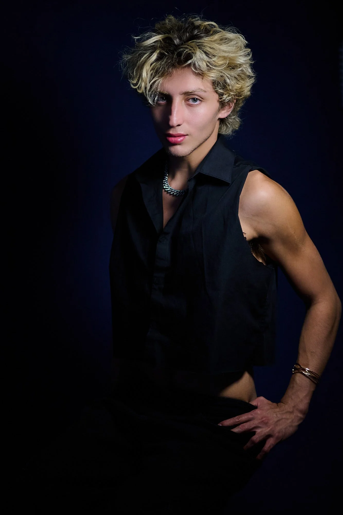 Person with blonde, curly hair wearing a black sleeveless shirt, silver necklace, and bracelets posing against a dark background.