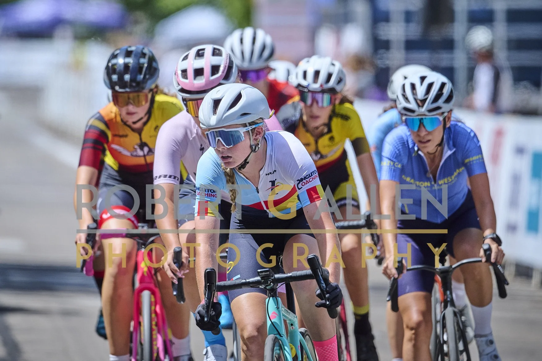 Group of female cyclists wearing helmets and colorful jerseys racing on a city street.