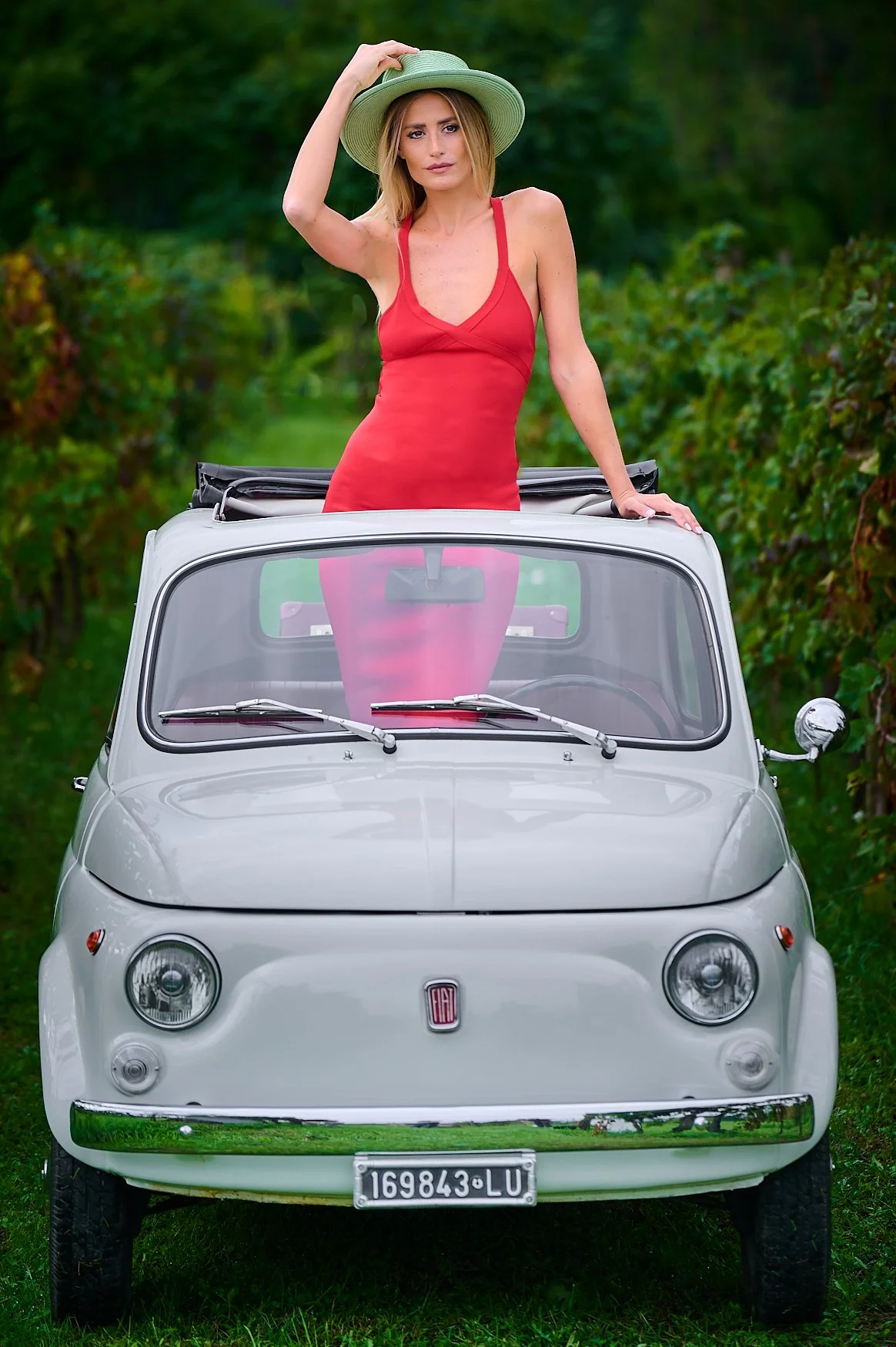 A woman in a red dress and wide-brimmed hat standing up through the roof of a small white vintage Fiat car in a green vineyard.