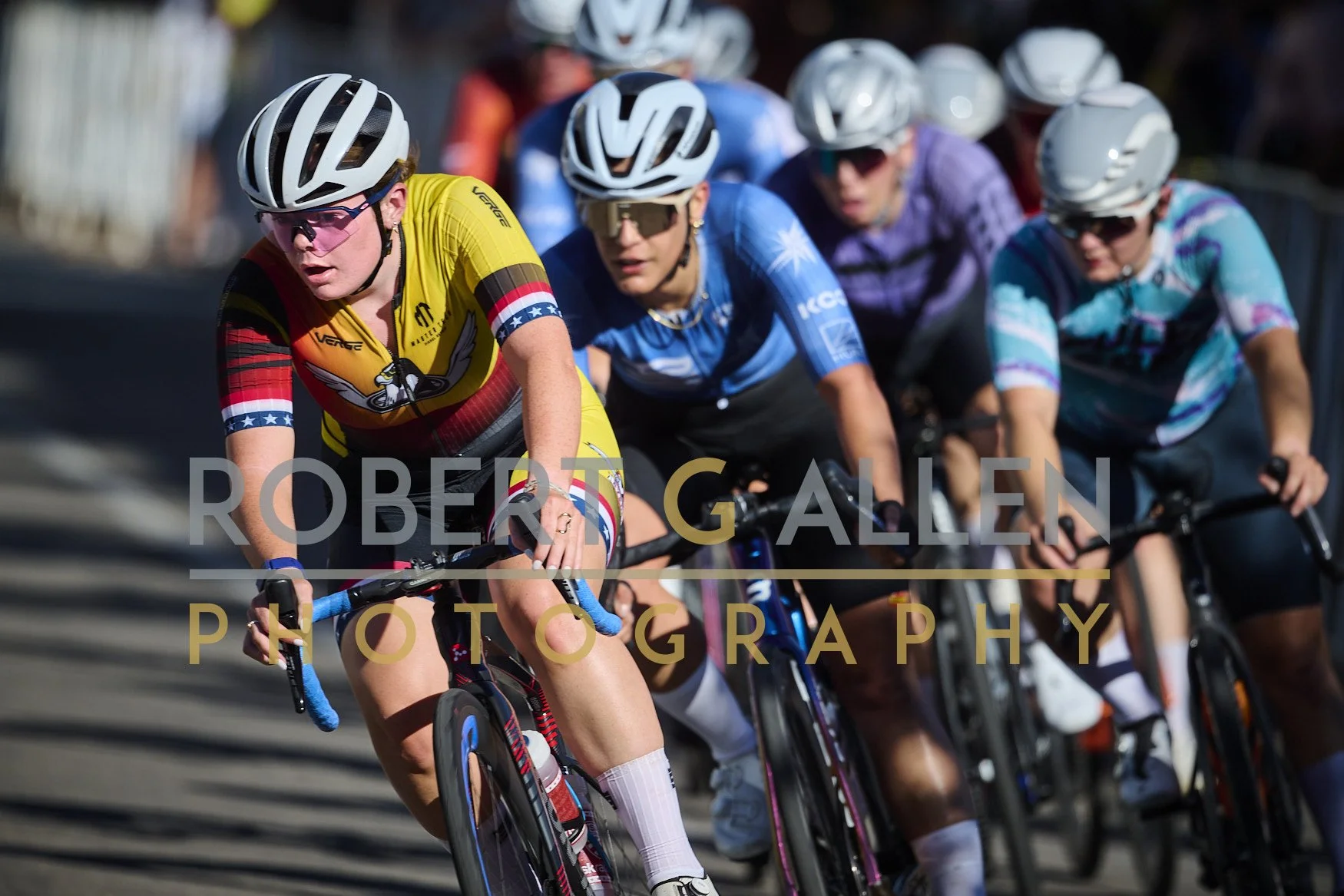 Cyclists racing in a group, wearing colorful jerseys and helmets, riding on a sunny day.