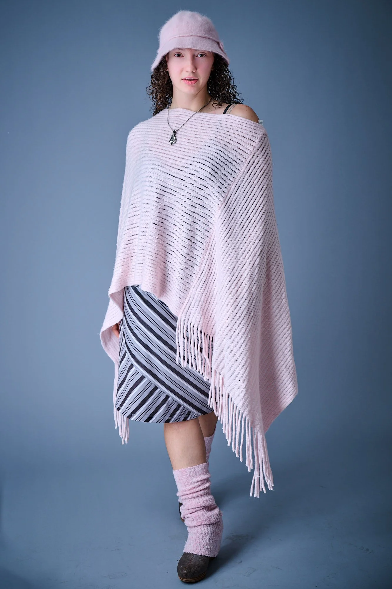 A young woman with curly hair wearing a pink hat, a black strap tank top, a pink oversized knit sweater, a gray and white striped skirt, pink knee-high socks, and brown shoes, standing against a blue background.