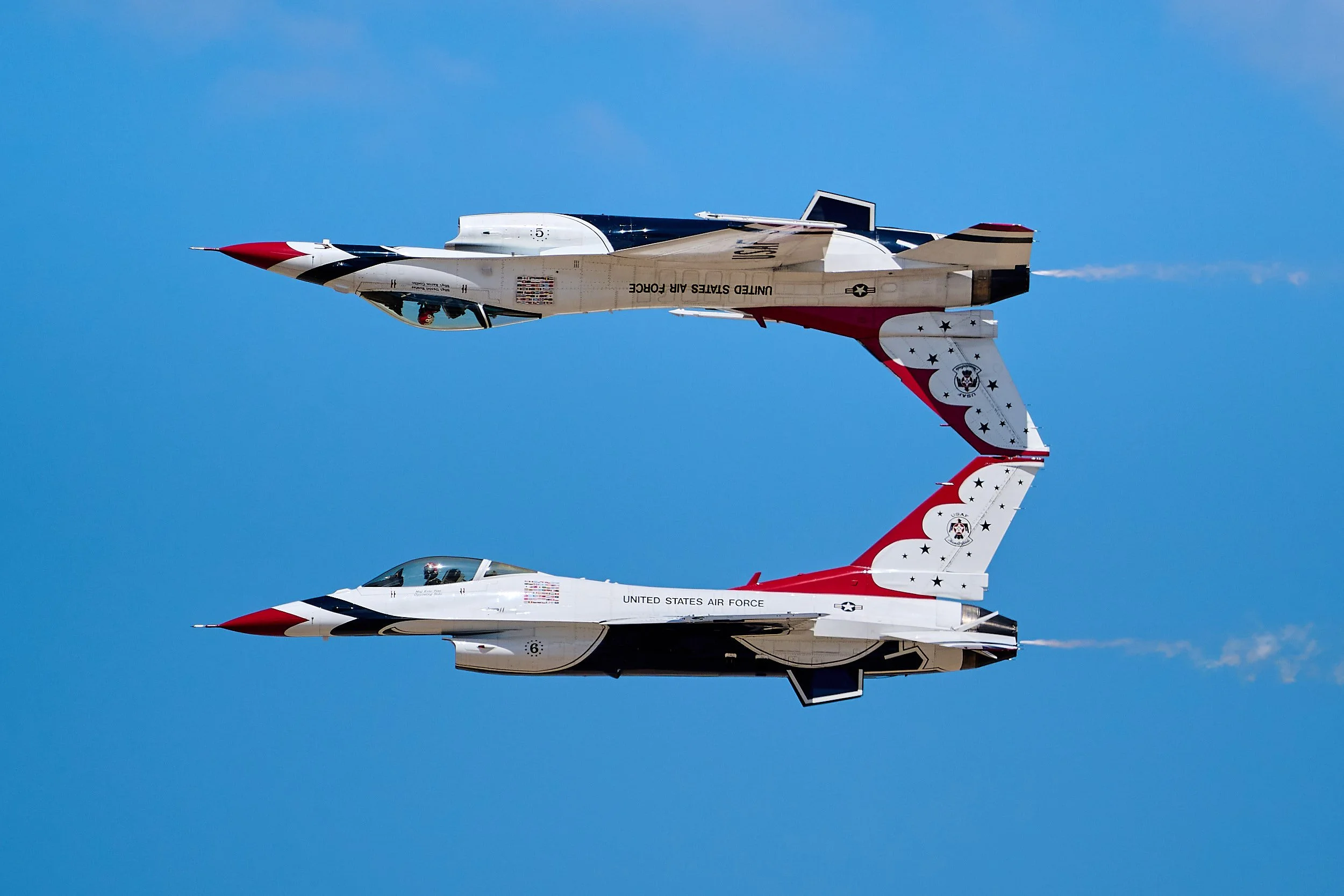 Two United States Air Force fighter jets flying in formation against a clear blue sky, with the top jet painted in patriotic red, white, and blue colors and the bottom jet in white with a red, white, and blue nose.