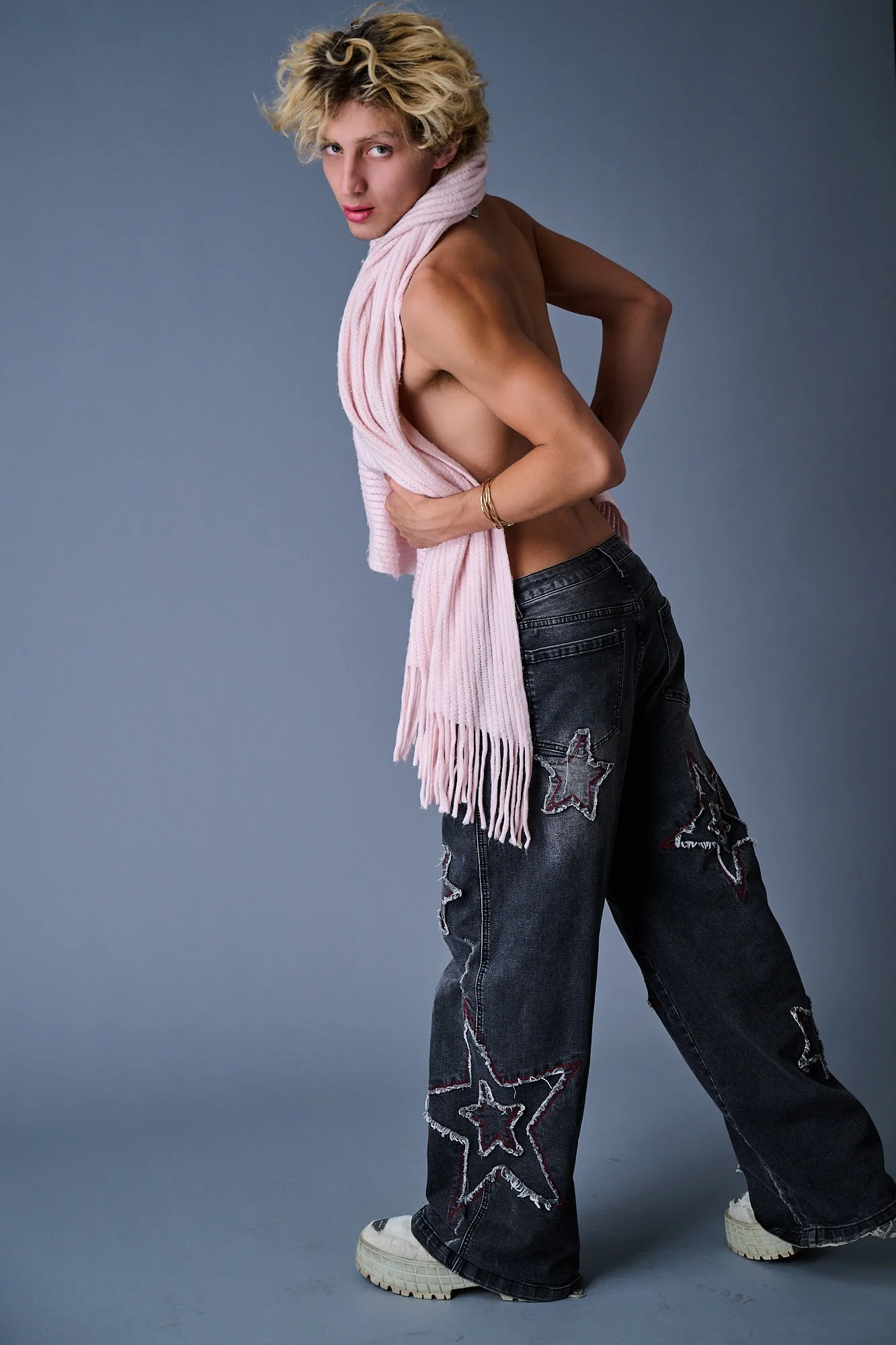 A person posing against a gray background, wearing baggy jeans with star patches, white sneakers, a pink scarf, and no shirt.