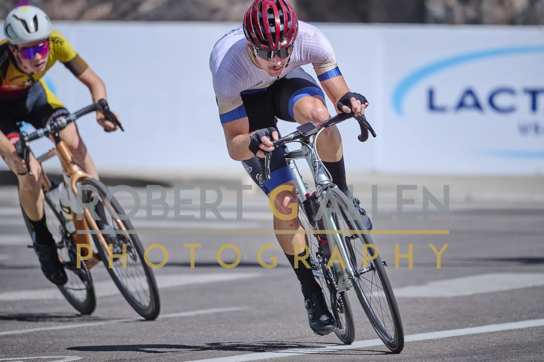 Two male cyclists racing on a road during daytime, wearing helmets and sports gear, leaning into a turn.