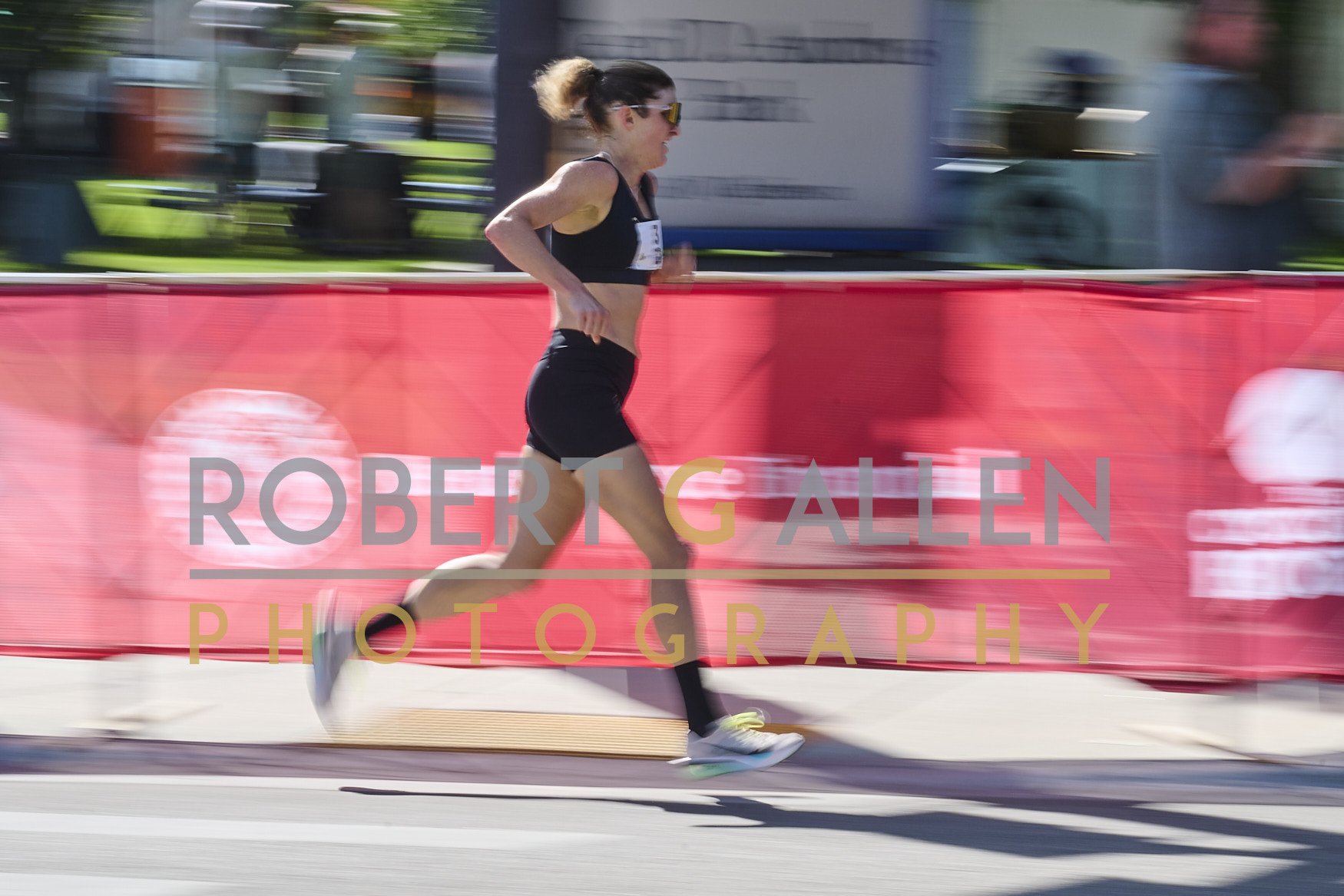 A female runner wearing black running clothes and sunglasses participating in a race, running past a red barrier with the words "Robert G. Allen Photography" on it, in an outdoor city setting.