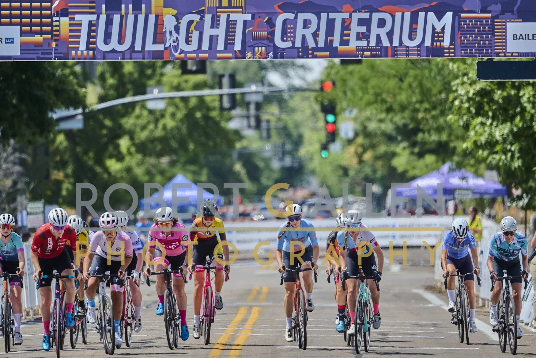 Group of female cyclists wearing helmets and colorful jerseys riding on a city street during a race, with trees lining the road and traffic lights overhead.