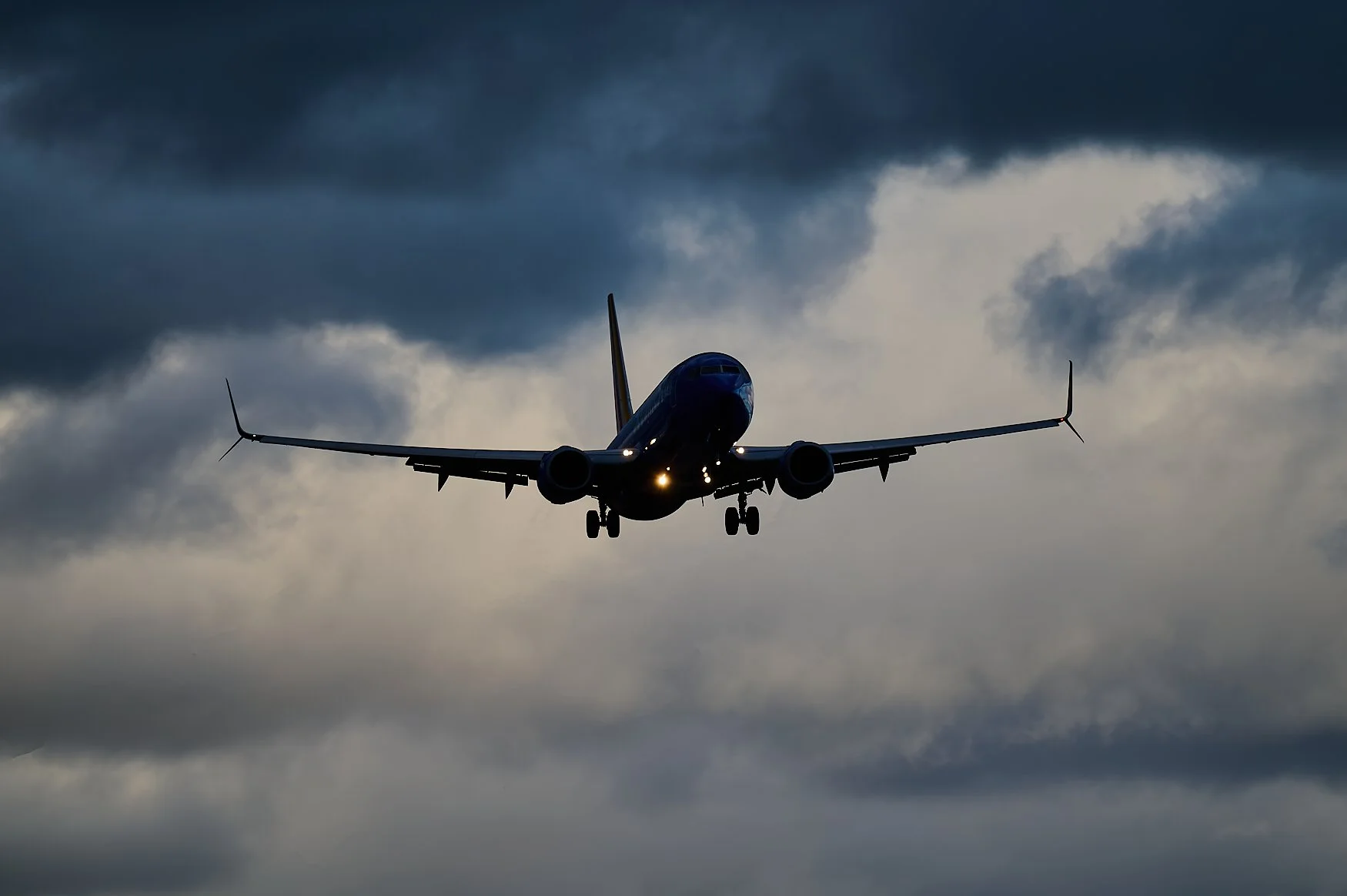 A commercial airplane flying through a cloudy sky, with dark clouds above and a break in the clouds showing lighter sky behind.