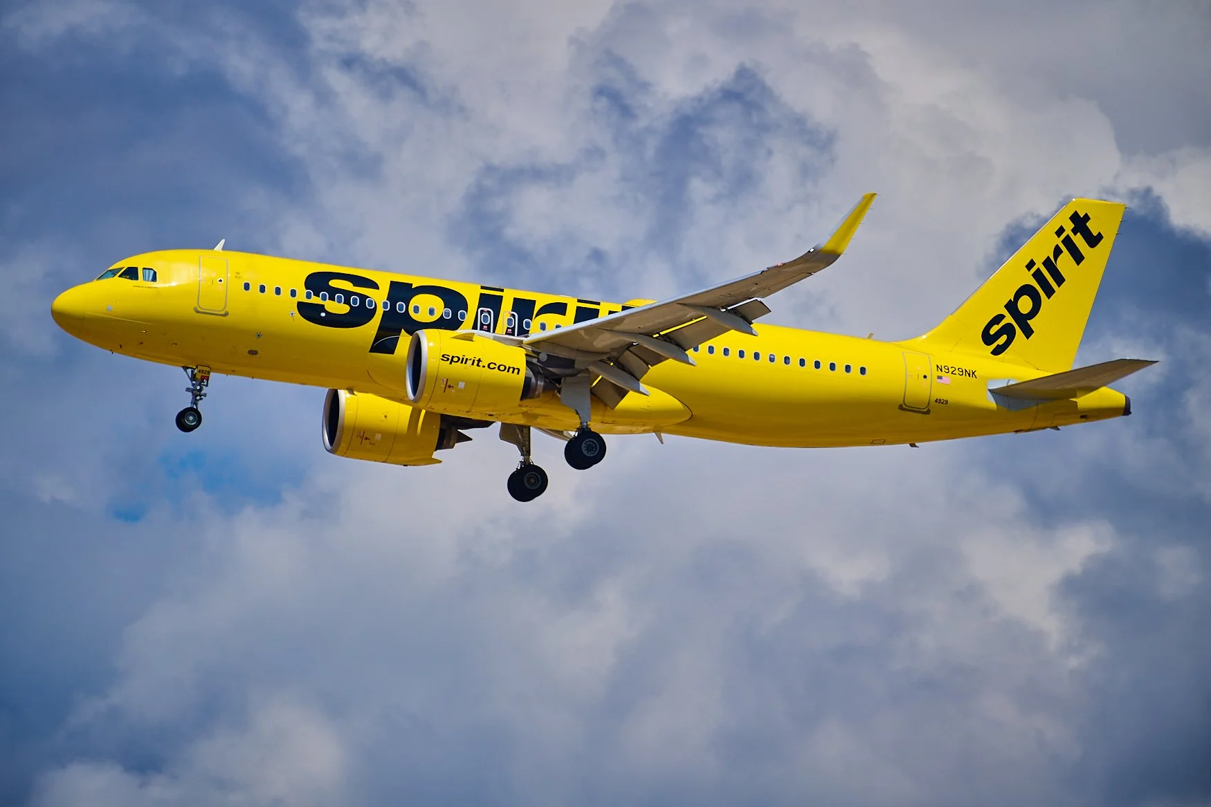 A bright yellow Spirit Airlines airplane flying through the sky with clouds in the background.