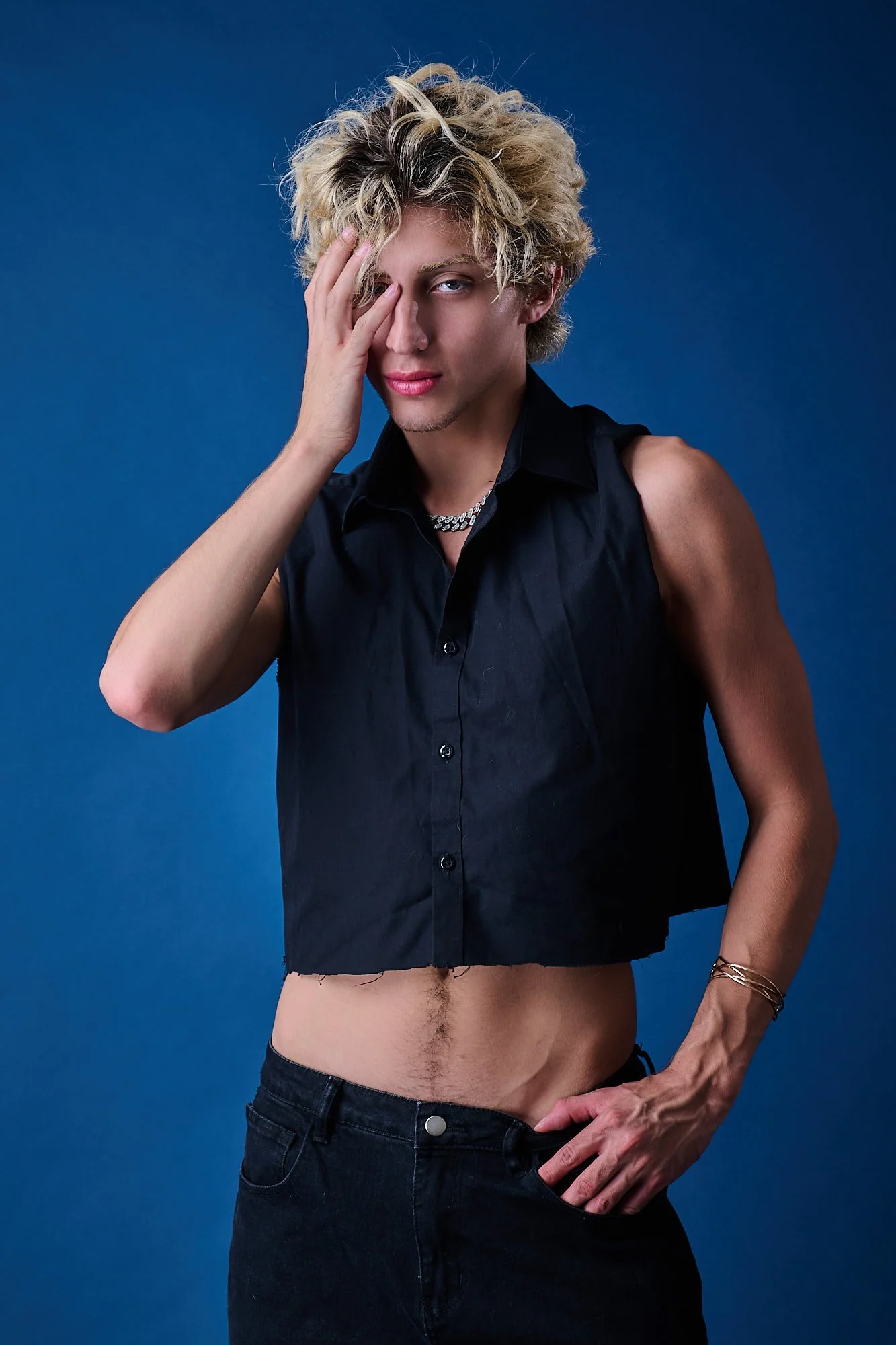 A young person with blonde, curly hair, wearing a sleeveless black shirt and black jeans, covering one eye with their hand, standing against a solid blue background.