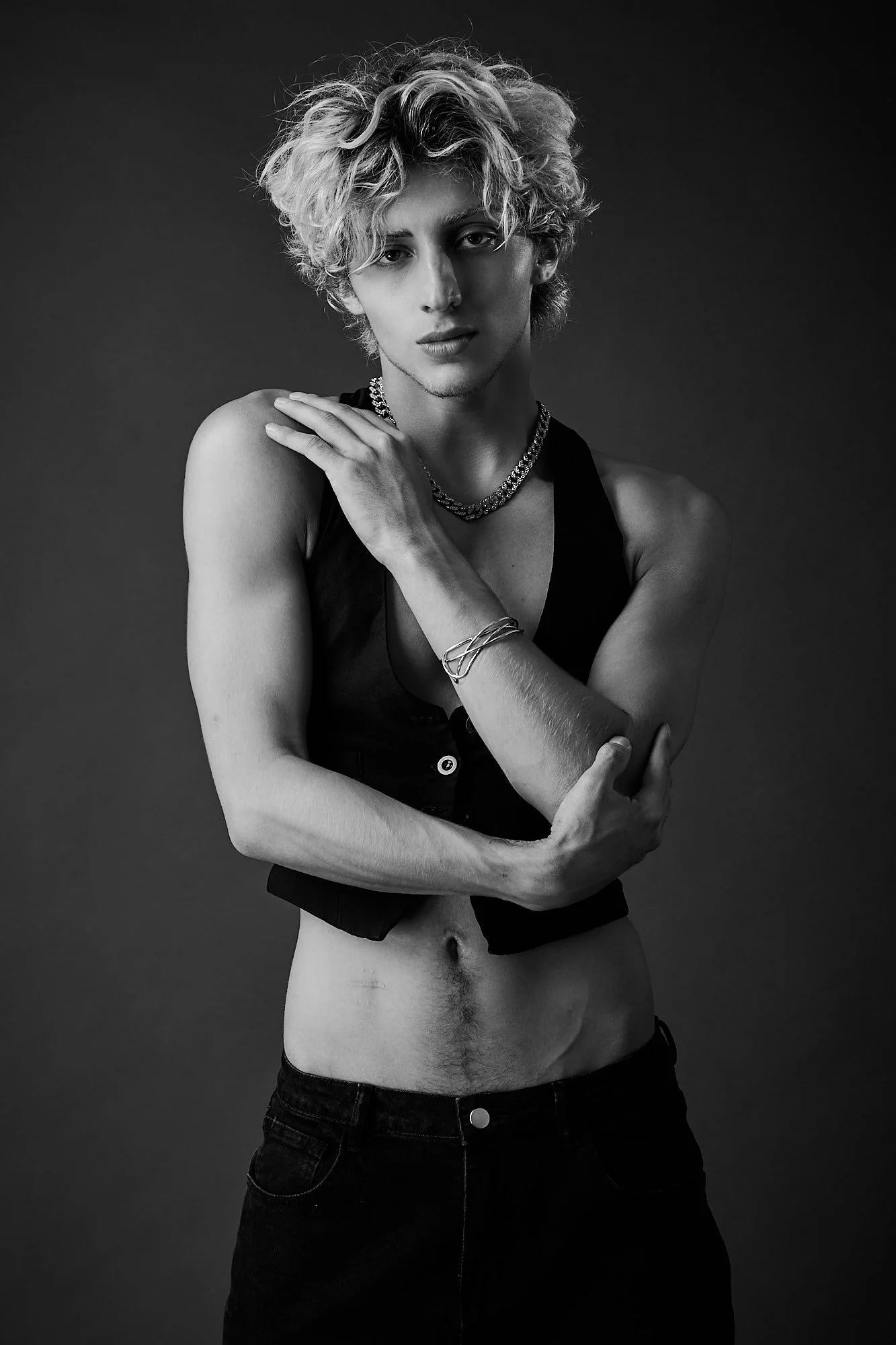Black and white portrait of a young woman with curly hair, wearing a sleeveless top, layered necklaces, and bracelets, posing against a plain dark background.