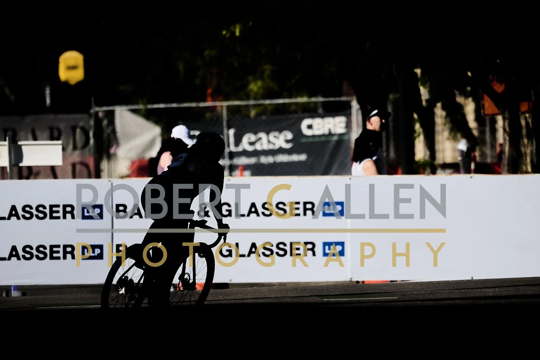 Silhouette of a person riding a bicycle in front of a large sign that reads "Babich & Glasen Photography" at an outdoor event, with other people and trees in the background