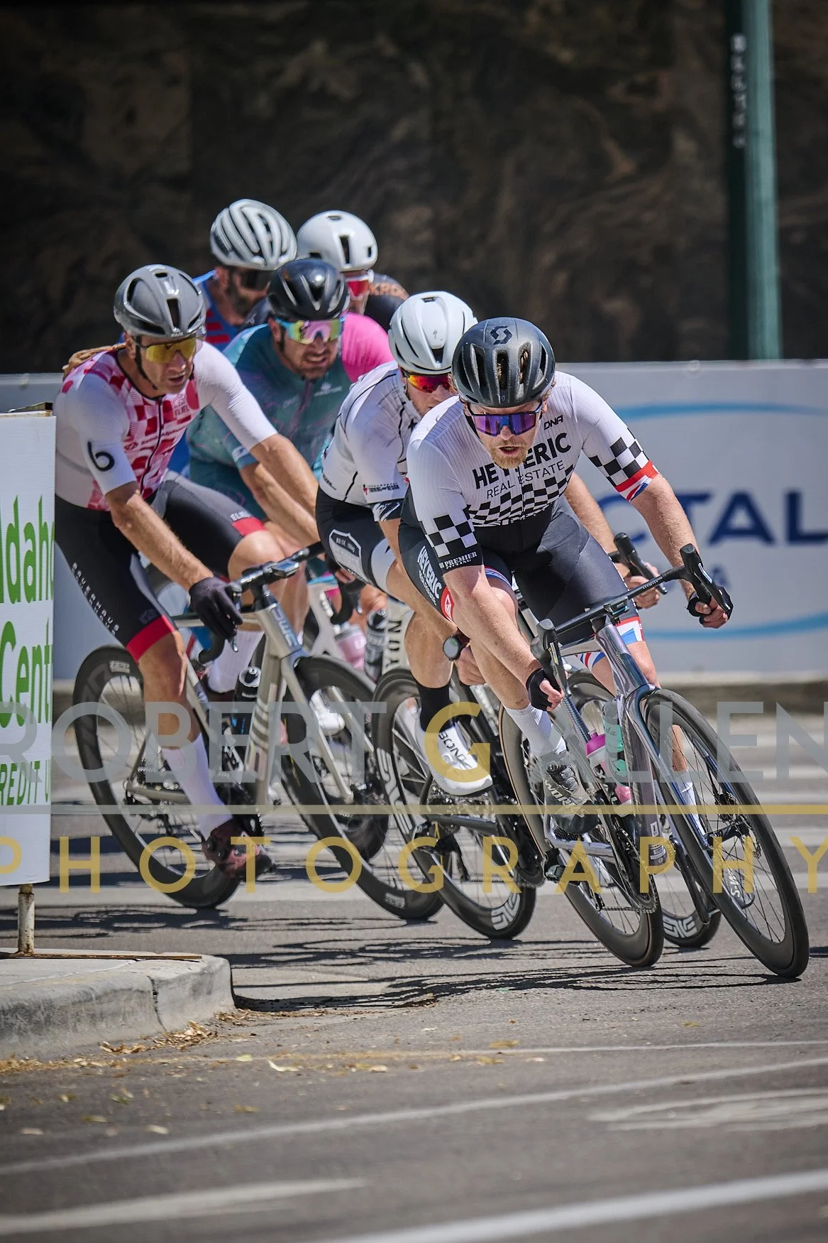 Group of cyclists racing closely on a city street, leaning into a turn. They are wearing helmets, sunglasses, and colorful cycling jerseys.