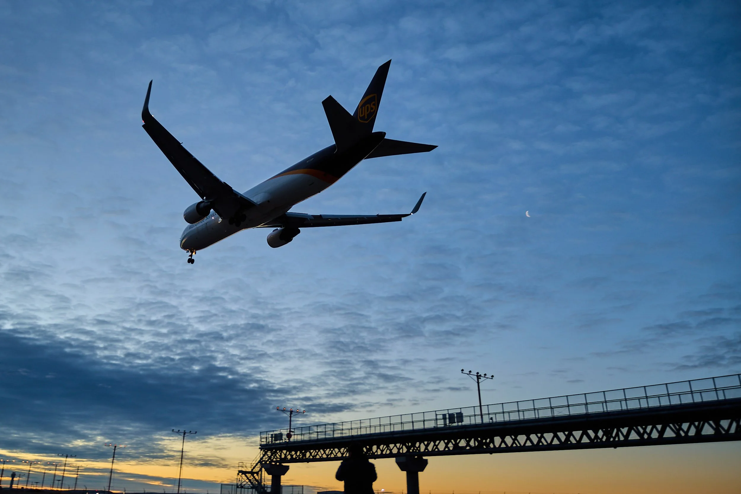 An aircraft with UPS logo flying in the sky during sunset or sunrise, with a silhouette of a person near an airport or airfield infrastructure and antennas below.