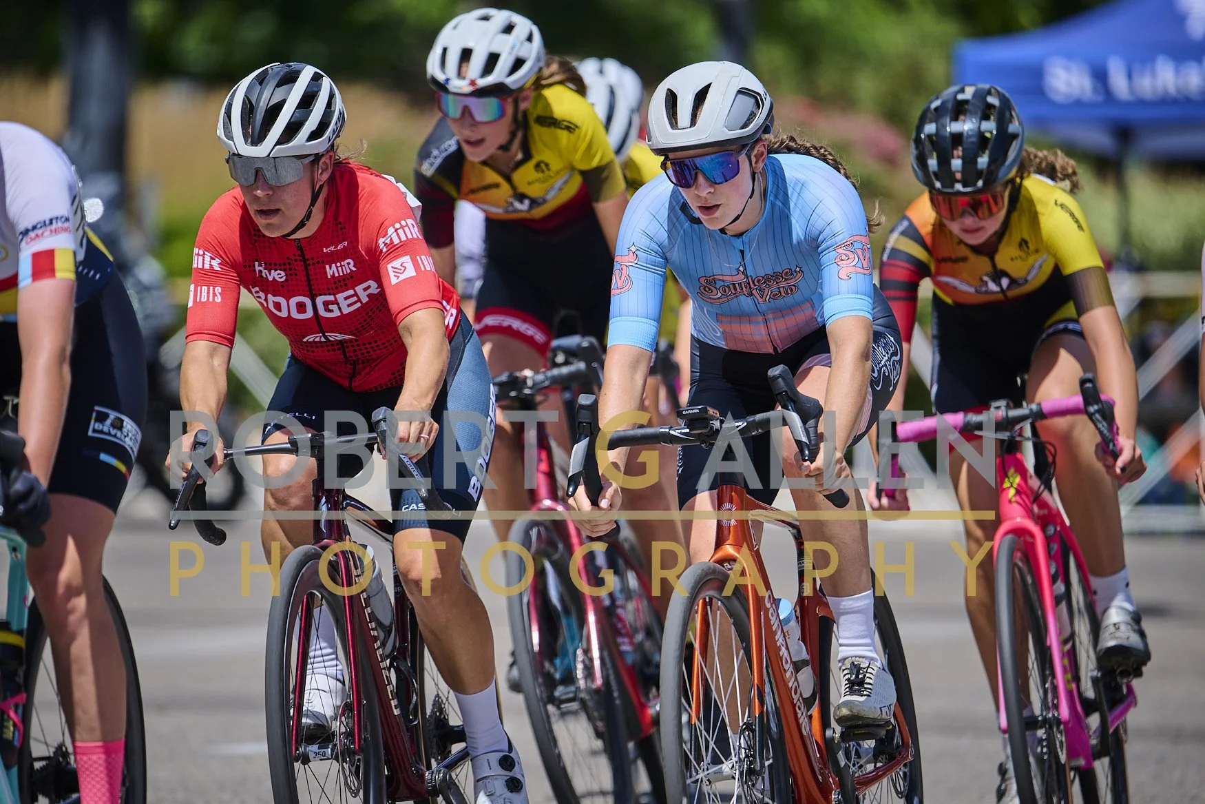 Cyclists participating in a race, wearing colorful jerseys and helmets, riding on a paved road with trees and a tent in the background.