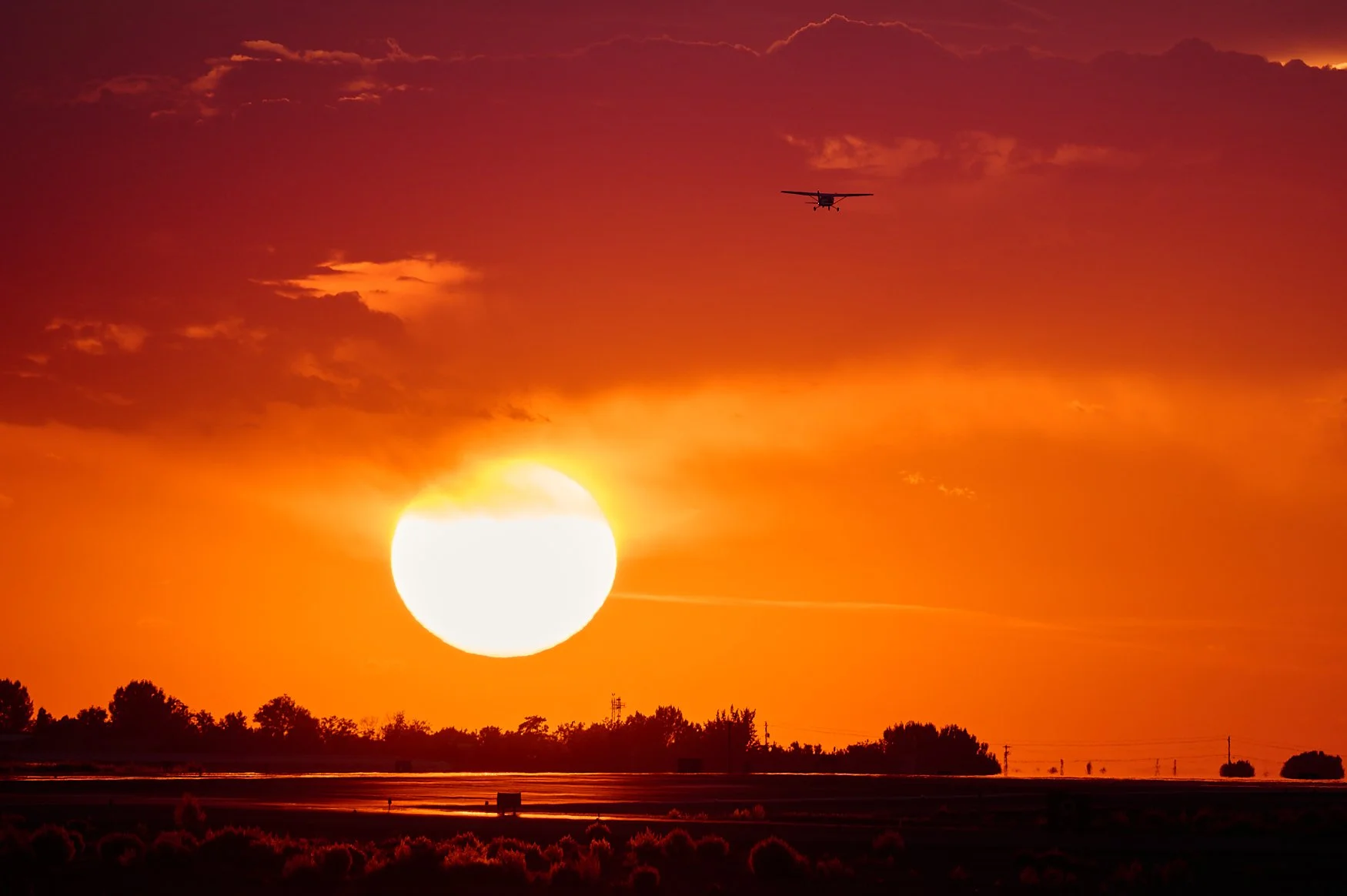Sunset with a large, bright sun near the horizon, orange sky with scattered dark clouds, silhouette of trees and power lines, and an airplane flying across the sky.