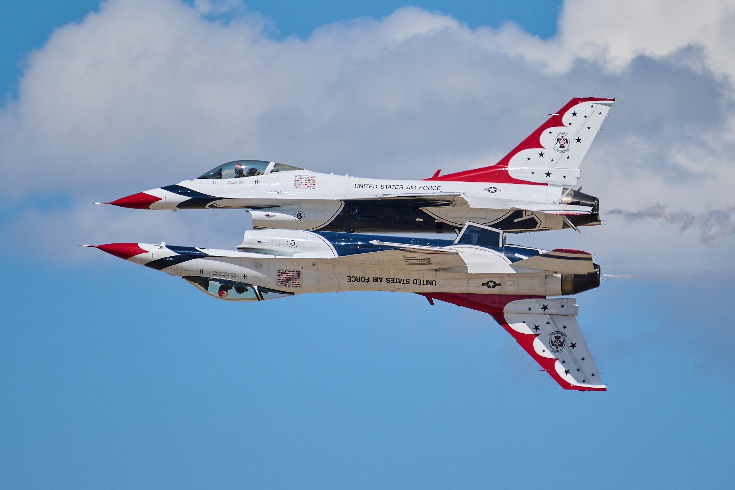An F-16 fighter jet in mid-flight against a partly cloudy blue sky, with a reflection showing on a shiny surface below.