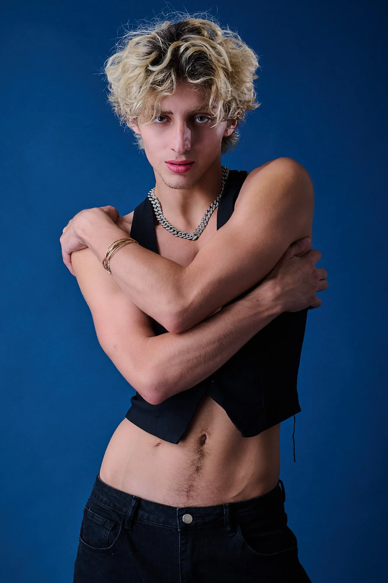 A young man with curly blonde hair and light skin is posing against a blue background. He is topless, wearing black shorts, and is accessorized with a silver chain necklace, gold bracelets, and a black sleeveless top. He is crossing his arms over his