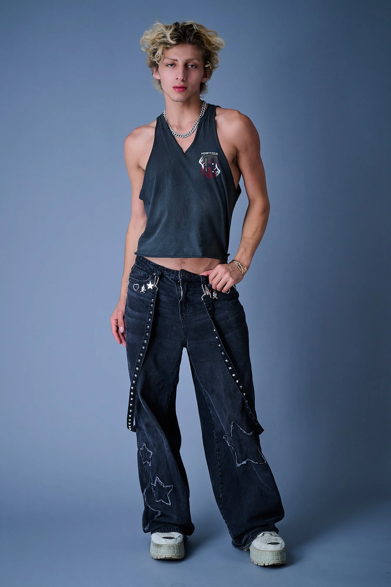 Young person with curly blonde hair wearing a black sleeveless shirt with a logo, baggy black jeans with star and heart patches, white sneakers, a silver chain necklace, and gold bracelets, standing against a blue-gray background.