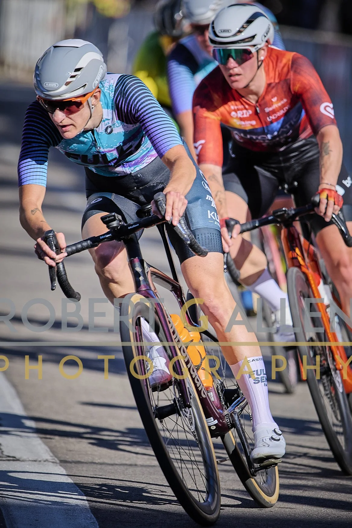 Cyclists riding a race on a sunny street, wearing helmets and colorful jerseys, with one cyclist in the foreground bending forward on a bike.
