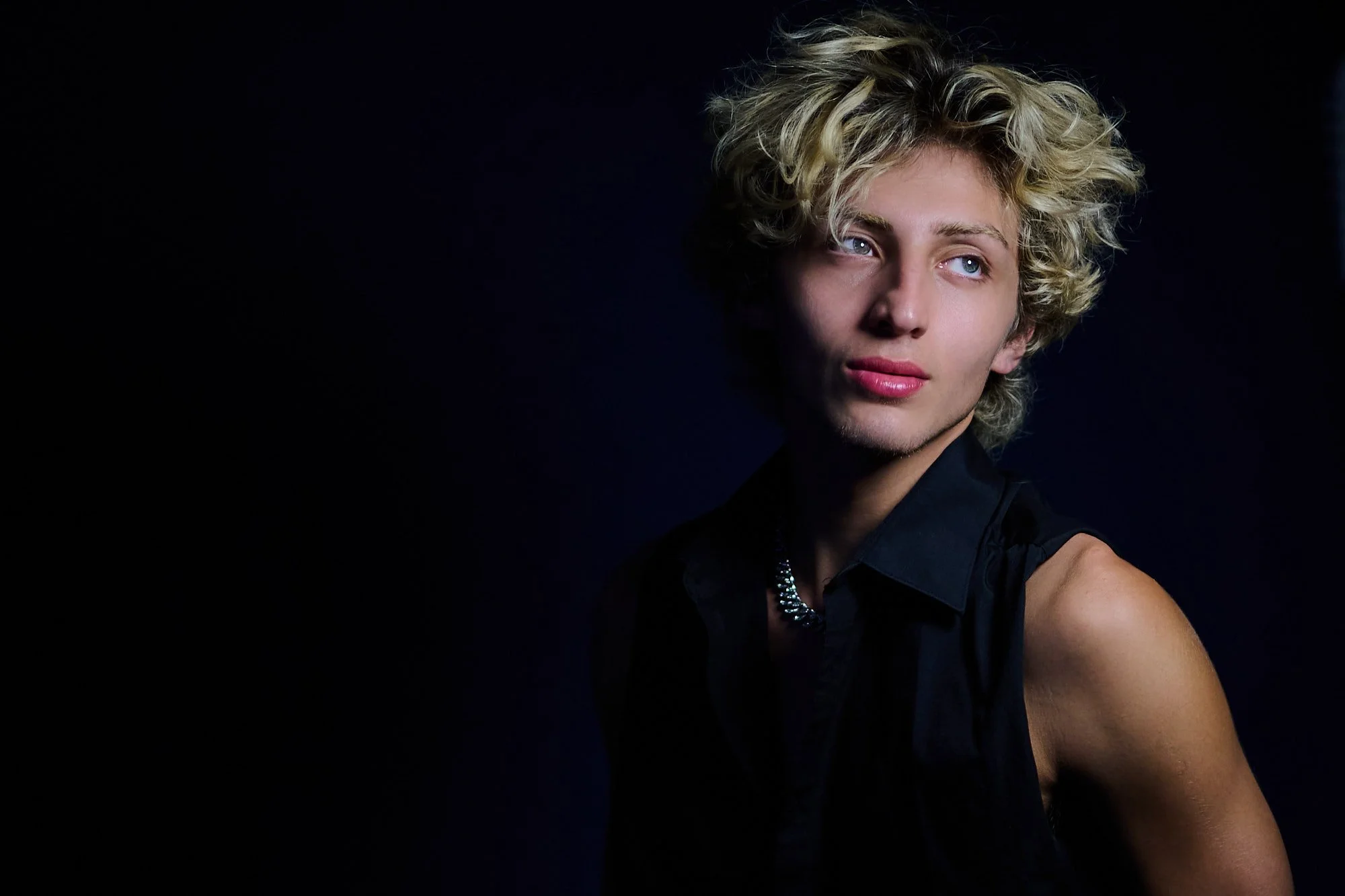 A young person with curly, blonde hair and blue eyes, wearing a black sleeveless top and a silver necklace, against a dark background.