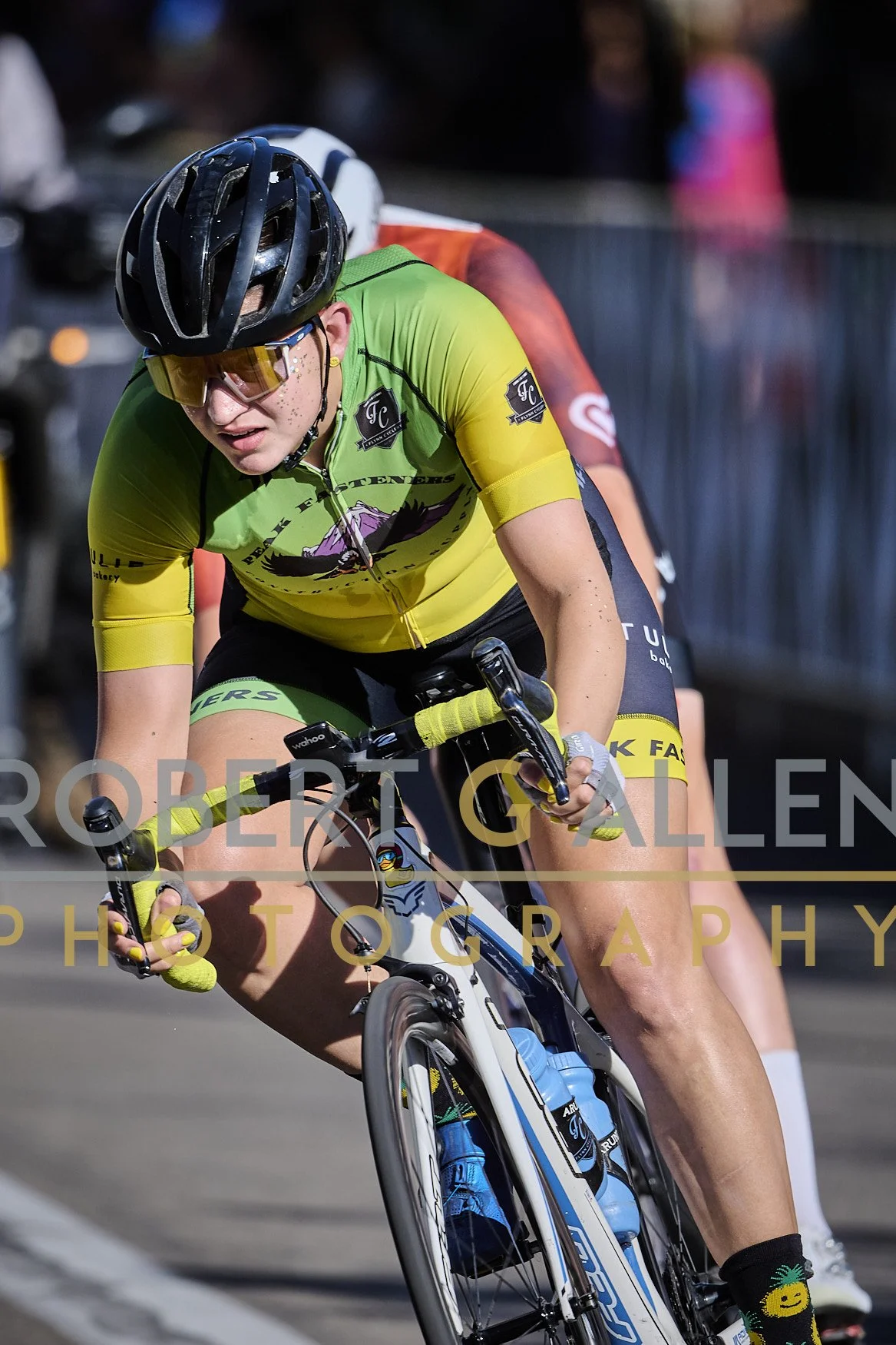 A cyclist in a green and yellow kit riding a racing bicycle during a race, wearing a black helmet and yellow-tinted glasses.