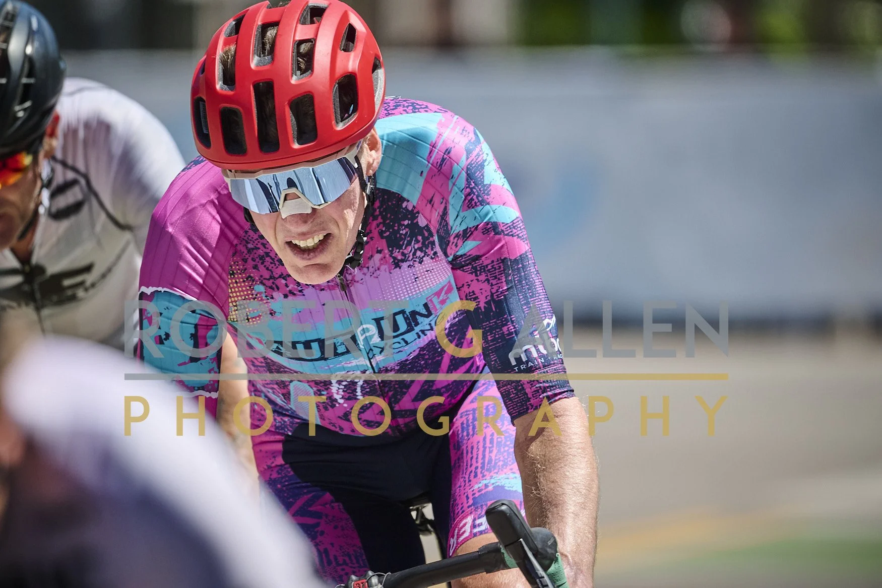 A man wearing a pink and blue cycling jersey, red helmet, and reflective sunglasses, riding a bicycle during a race or ride.