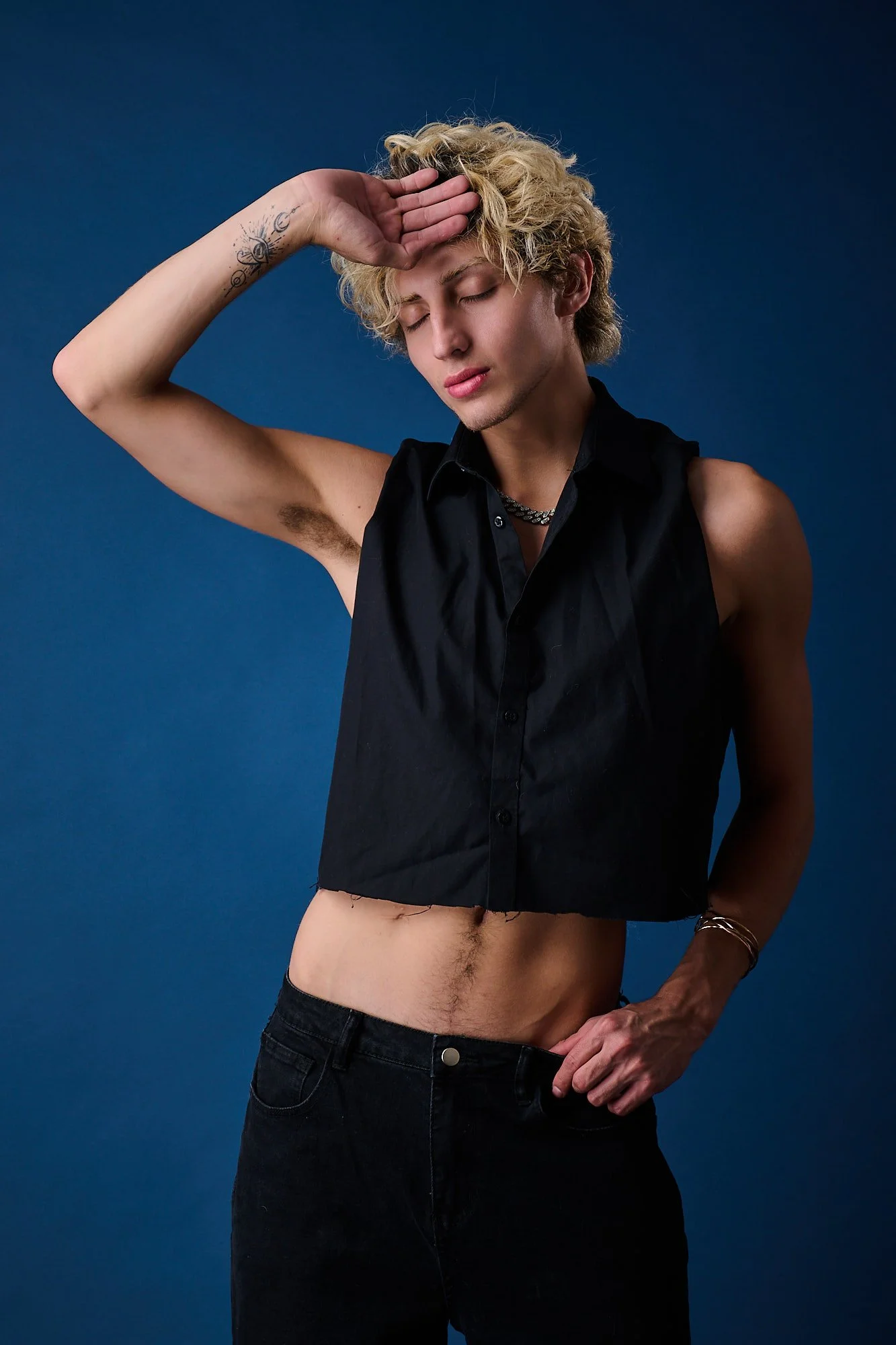 A young man with blonde curly hair, wearing a sleeveless black shirt, black jeans, and a chain necklace, standing against a blue background with one hand on his forehead and eyes closed.