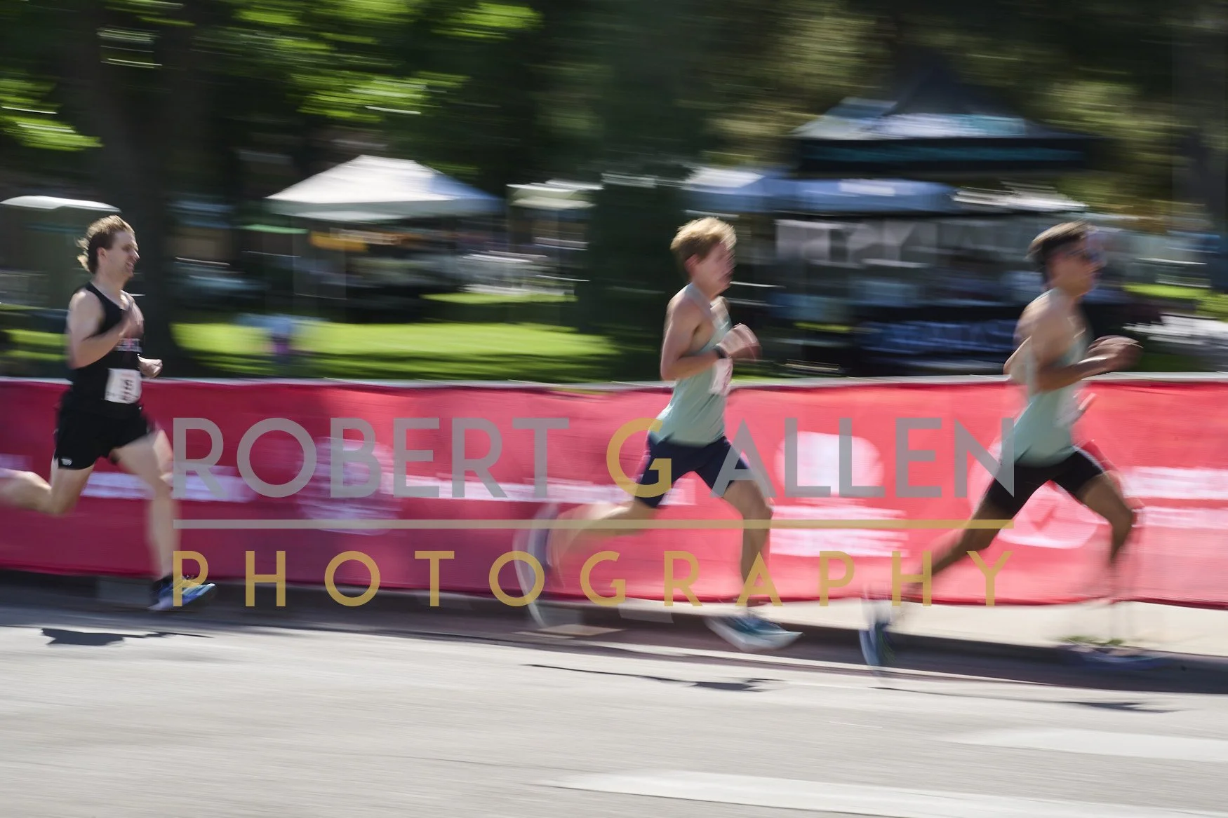 Three runners in a race on a sunny day, running along a street with trees and tents in the background, and a pink banner with 'Robert Gallen Photography' overlaid.