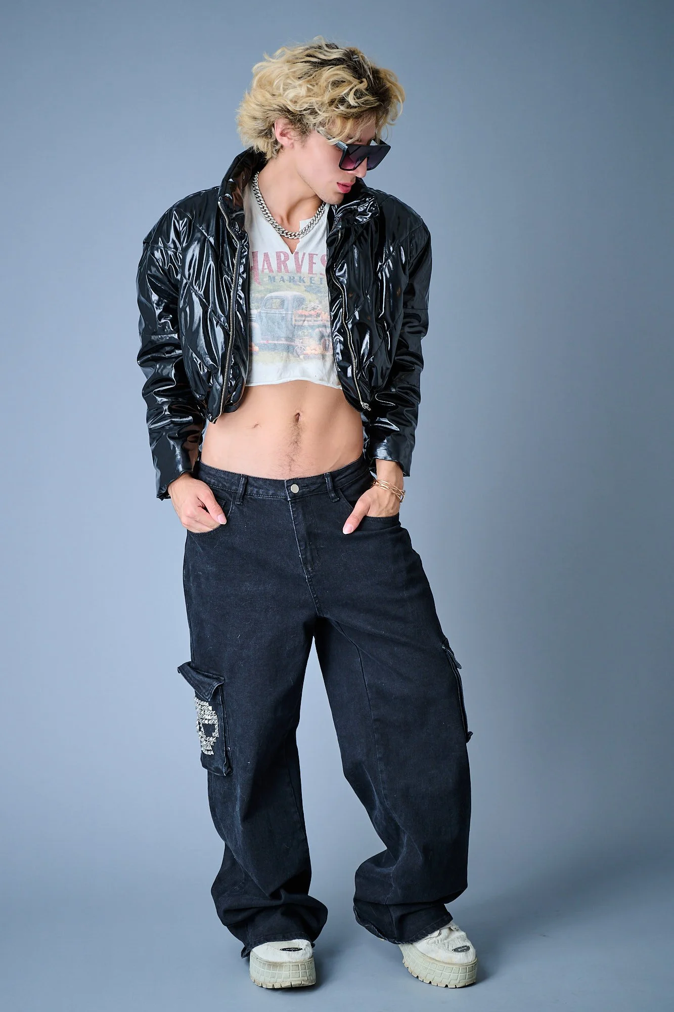 Person with curly blonde hair wearing large sunglasses, a shiny black jacket, a vintage 'Harvey' T-shirt, baggy black cargo pants, and white sneakers, standing against a plain gray background.