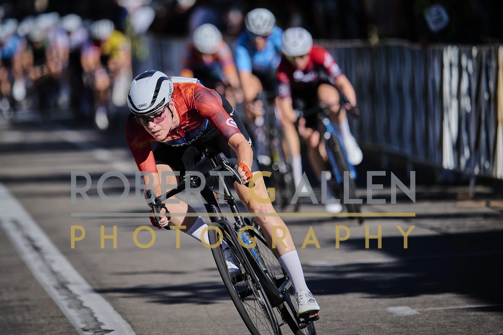 A cyclist in a red and black jersey wearing a white helmet and cycling sunglasses leans into a curve on a racetrack with a blurred group of cyclists behind him. The photo is watermarked with "Robert G. Allen Photography."