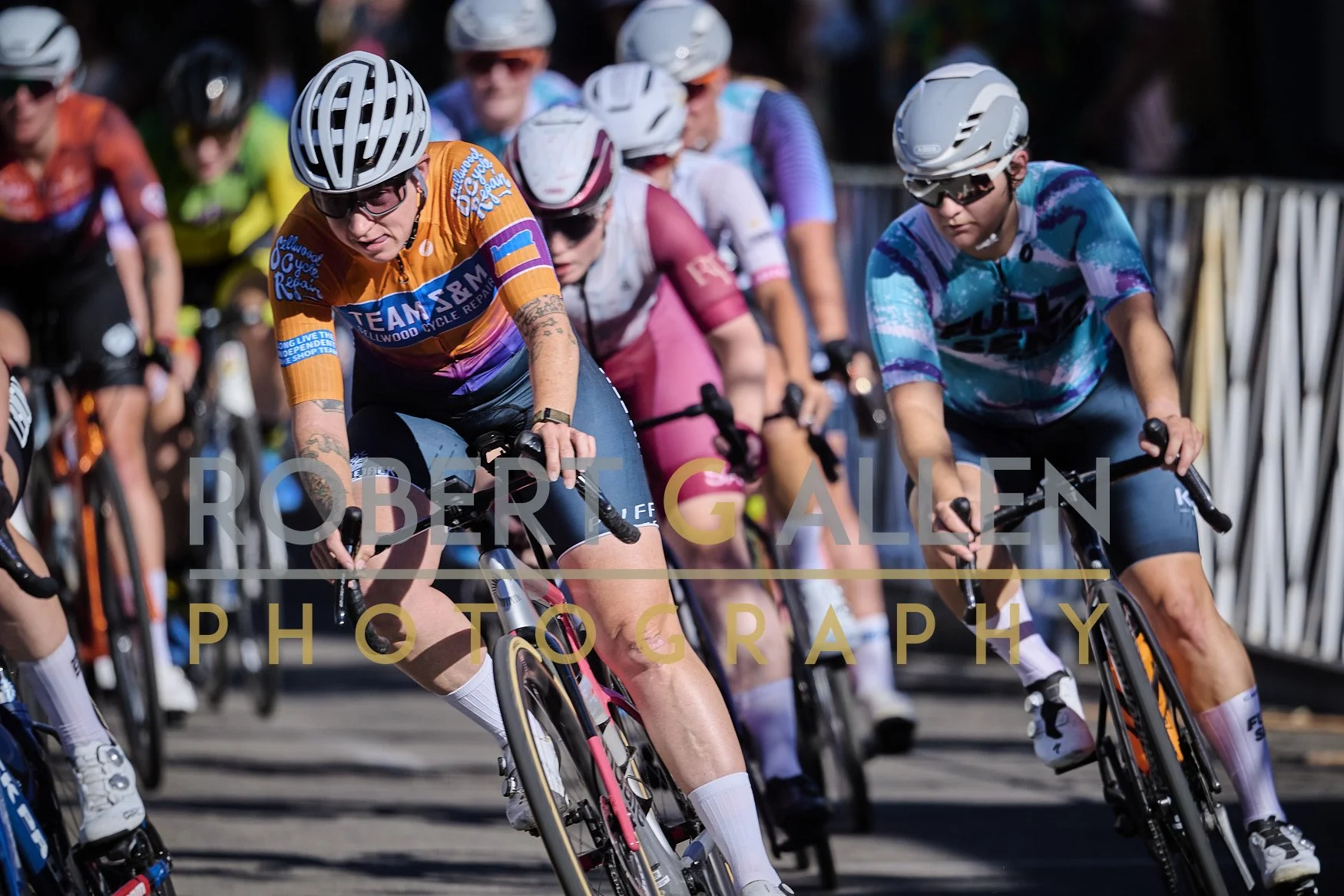 A group of cyclists racing, wearing helmets and colorful jerseys, closely packed together on a road.