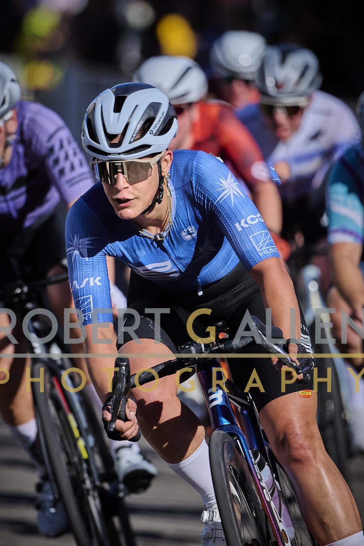 A group of cyclists riding together in a race, with the focus on a female cyclist wearing a blue jersey, white helmet, and sunglasses.