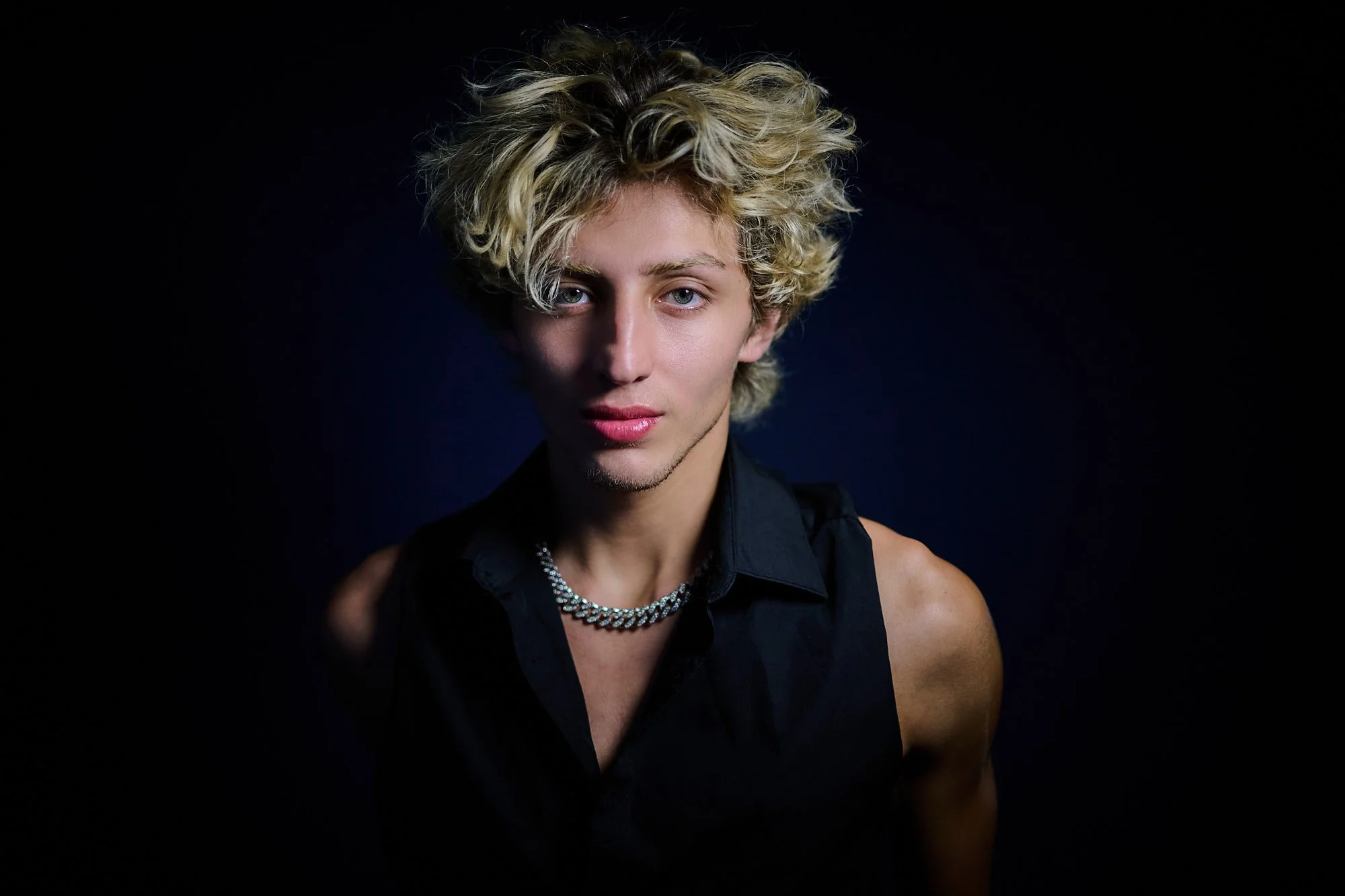 Close-up portrait of a young person with blonde, curly hair, blue eyes, wearing a black sleeveless top and a silver chain necklace, against a dark background.