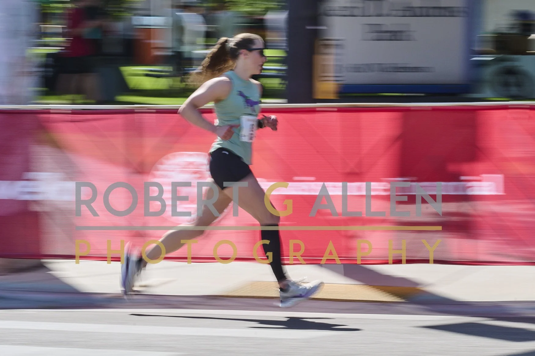 A female runner in athletic clothing runs along a street during a race, with a red barrier and signage in the background.