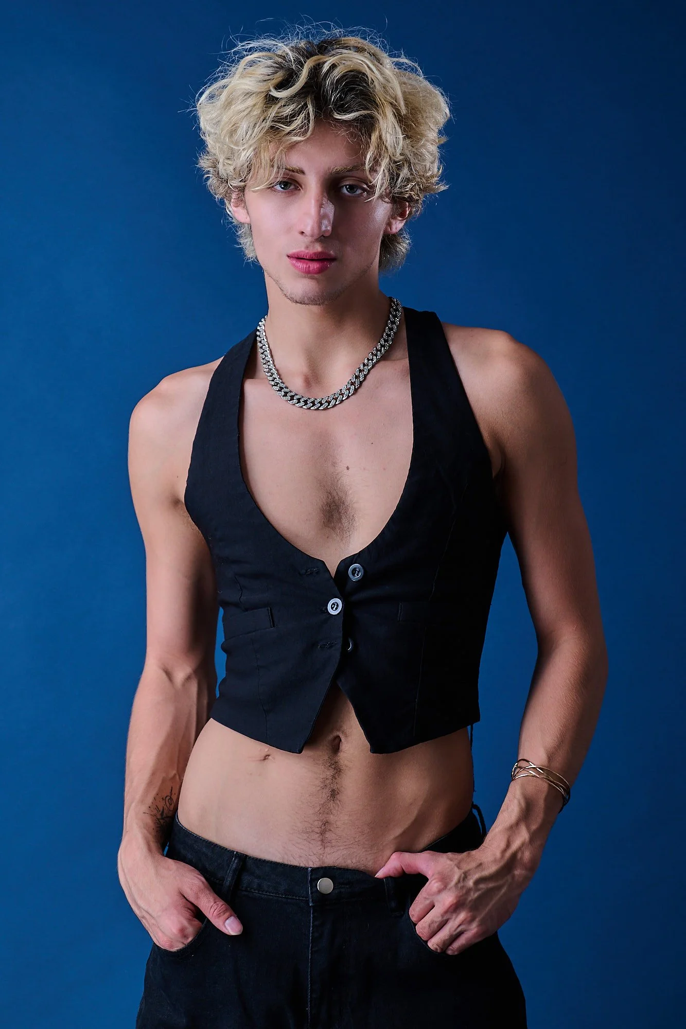 Young person with curly blonde hair wearing a black sleeveless vest, silver chain necklace, and black pants, standing against a dark blue background.