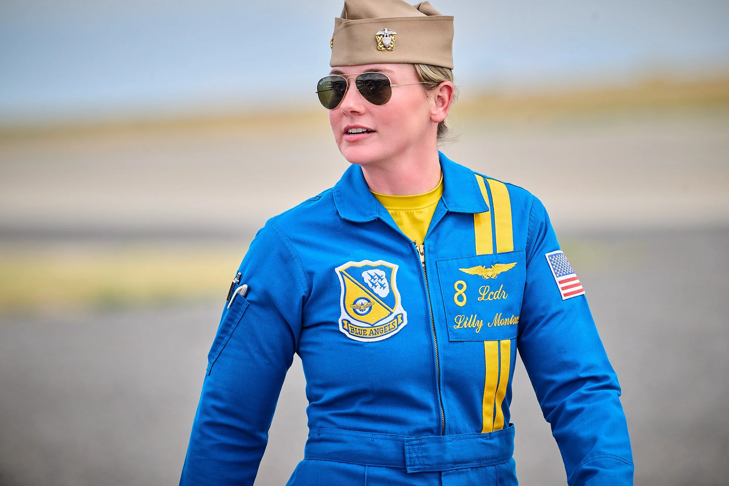 Woman in a blue flight suit with patches, wearing a tan flight cap with emblem, sunglasses, and a yellow undershirt, standing outdoors.
