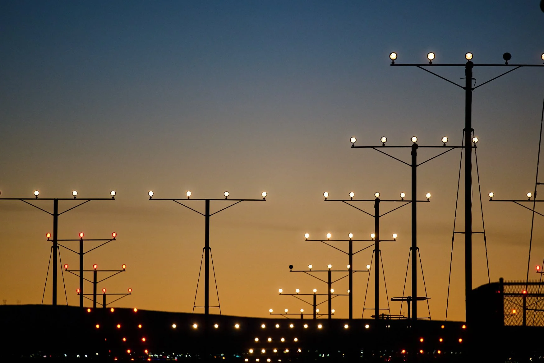 Multiple airport or runway lights on tall poles silhouetted against a sunset or dusk sky with a gradient of yellow, orange, and blue colors.