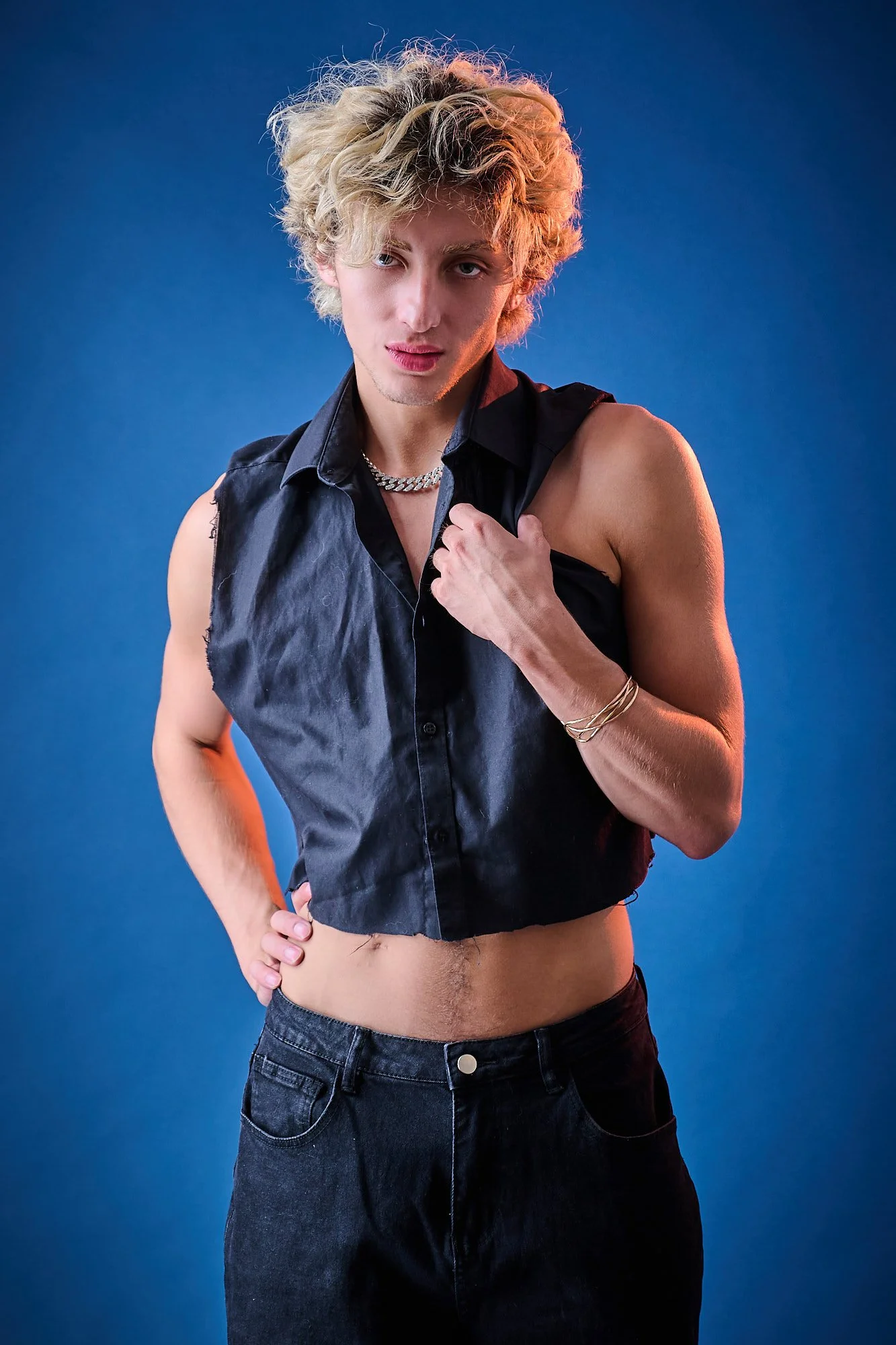 Young person with curly blonde hair wearing a sleeveless black shirt, dark jeans, jewelry, standing against a blue background.