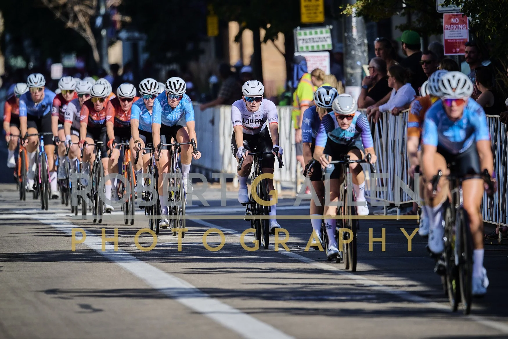 Cyclists wearing helmets and sports gear participating in a race on an urban street, with spectators behind barriers and green trees lining the street in the background.