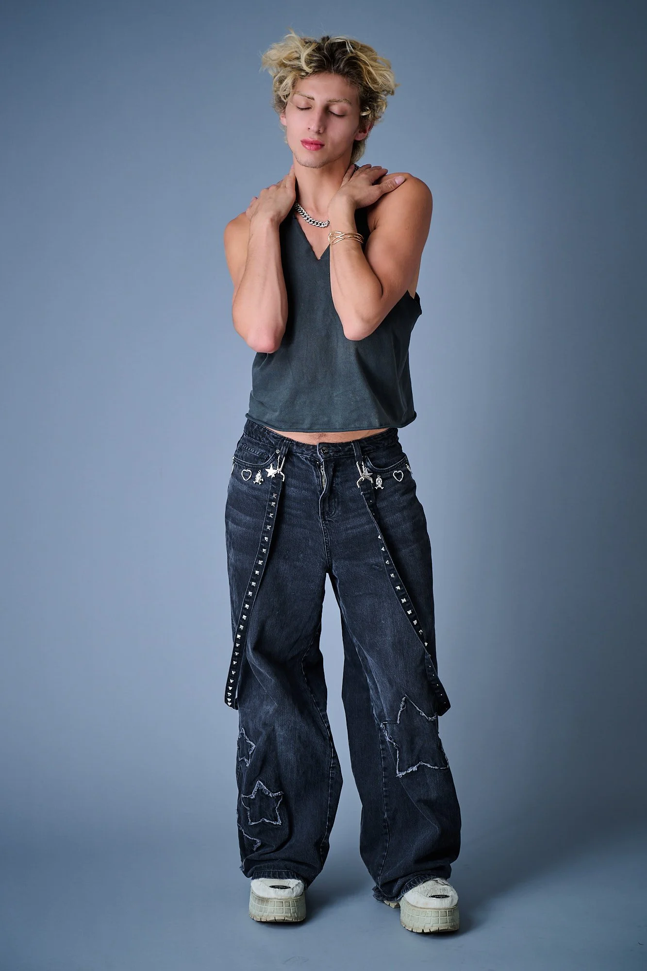 Young person with blonde, curly hair dressed in a black sleeveless top, baggy black jeans with star and heart patches, and white sneakers, standing against a gray background with eyes closed, touching their shoulders.