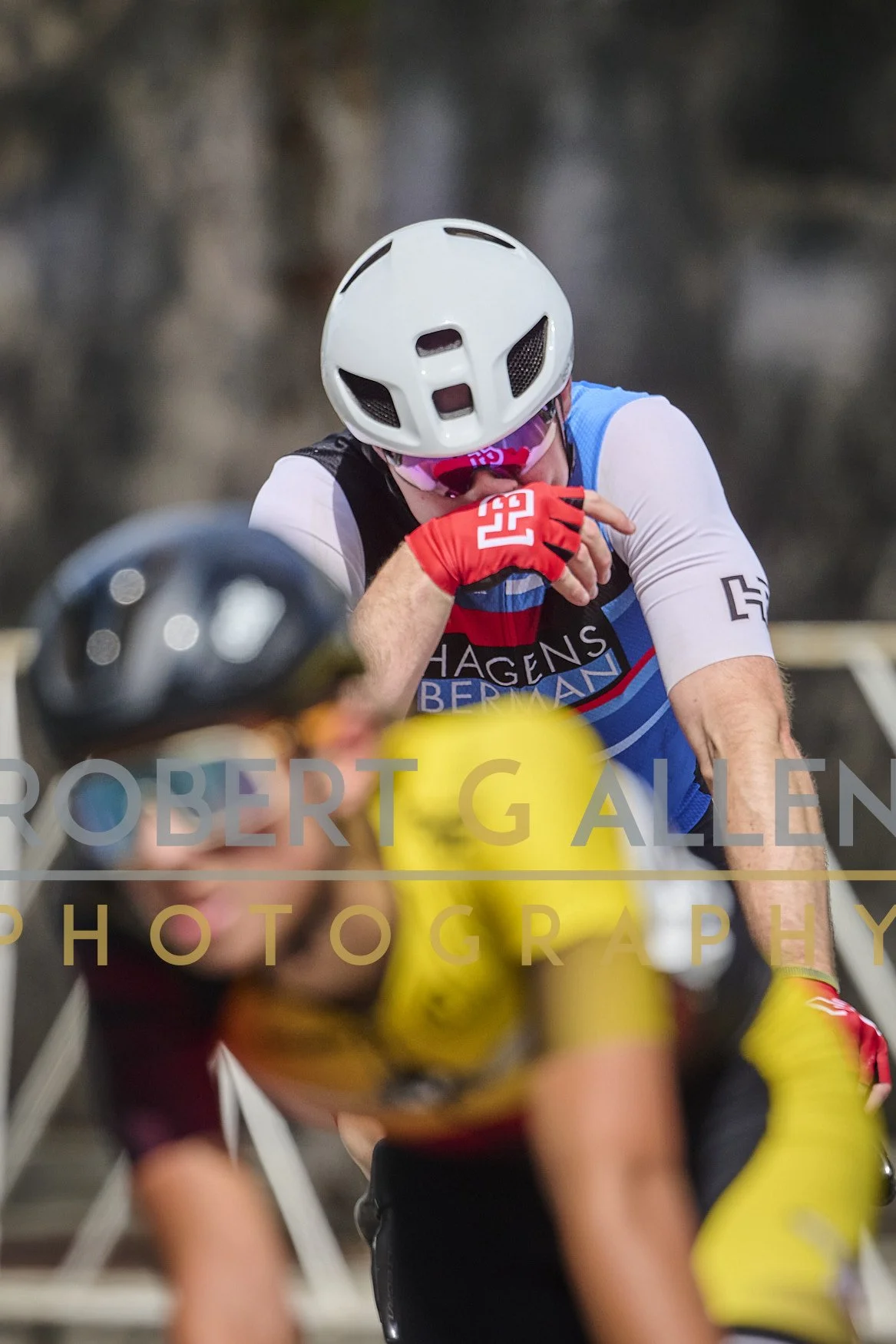 A cyclist wearing a white helmet, pink sunglasses, and a blue and black jersey, appears to be resting or catching a breath during a race. There is another cyclist in the foreground, slightly blurred, wearing a yellow jersey and black helmet.