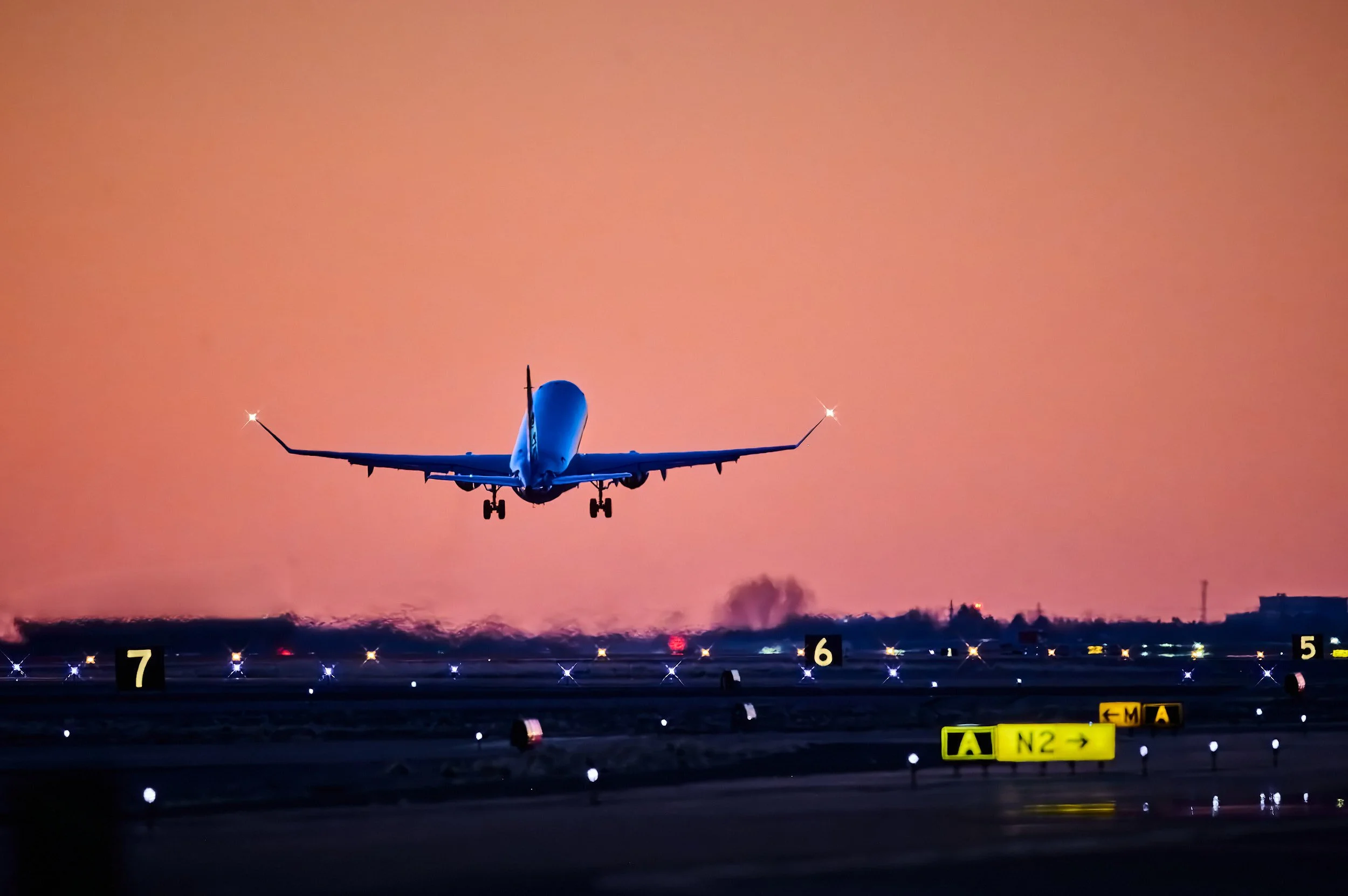 An airplane is landing on a runway during sunset with an orange sky in the background. The runway lights and signs are visible at the bottom of the image.