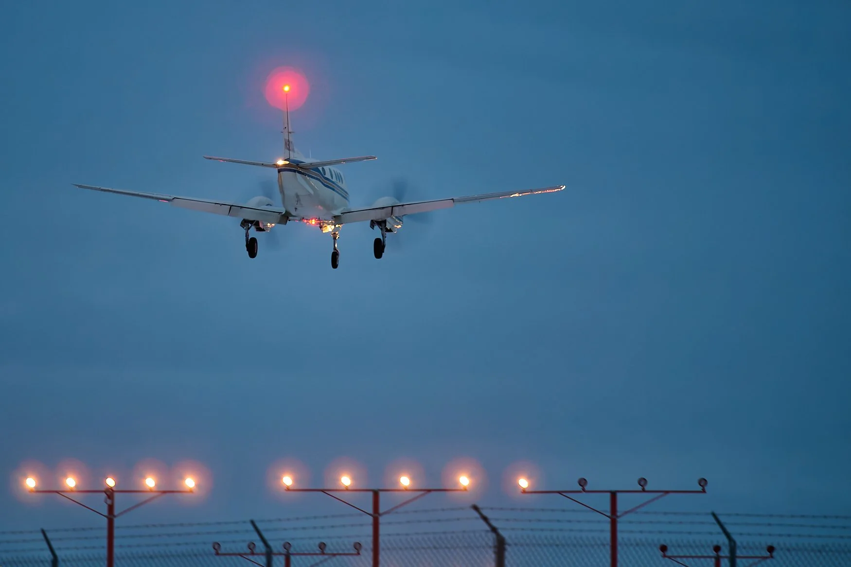 A small airplane landing on a runway at dusk with lights on the ground and a red light on top of the tail. The sky is cloudy and gray.